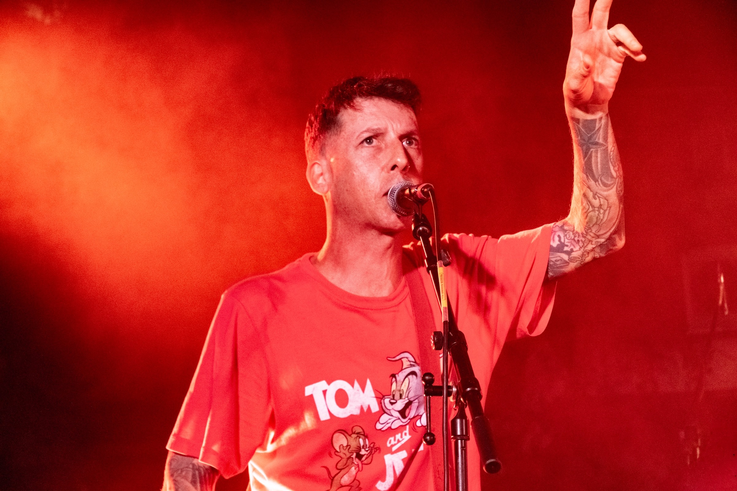 A tattooed singer in a Tom and Jerry t-shirt performs at a microphone bathed in red stage light, raising a peace sign