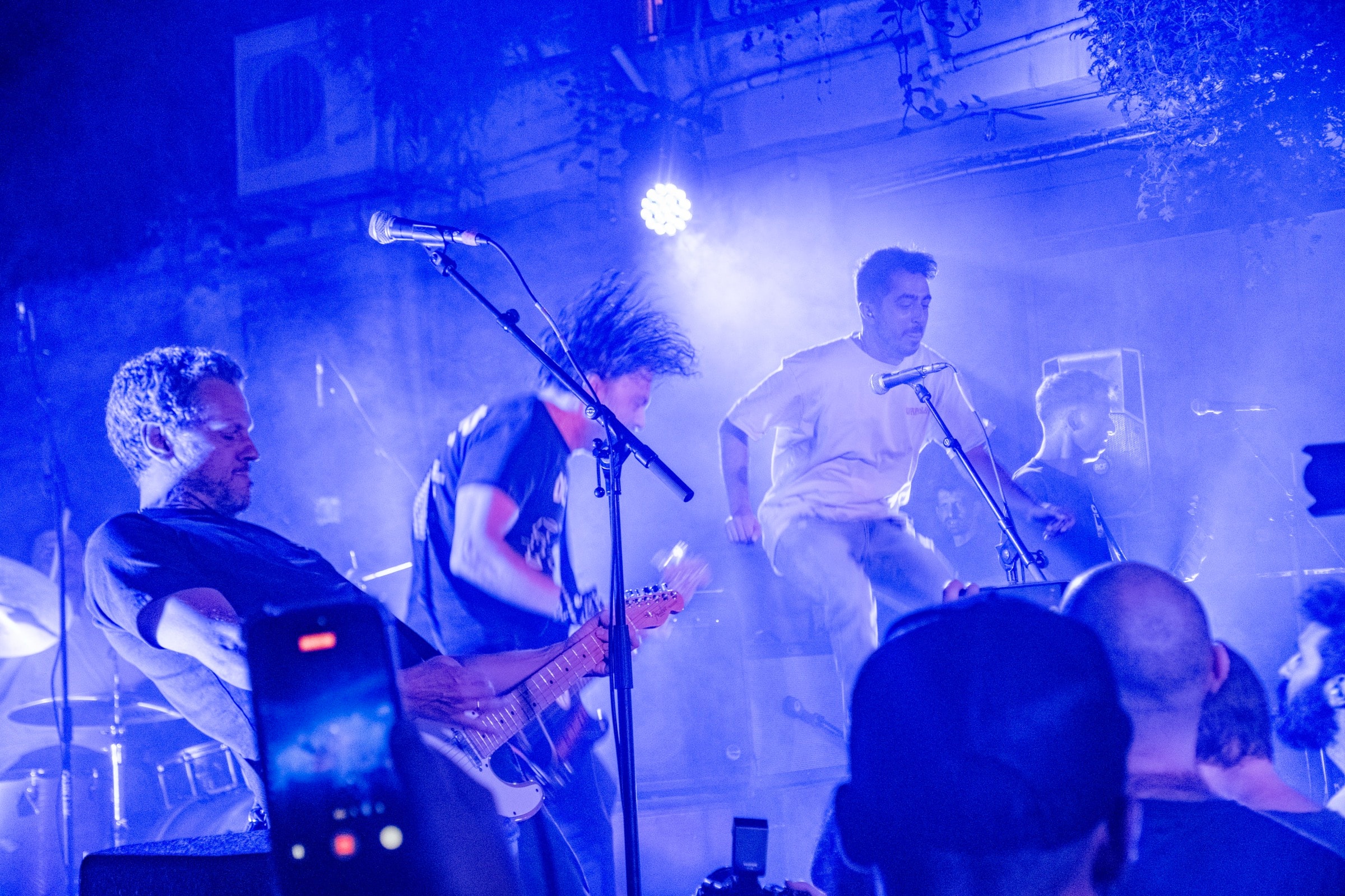 A band performs on an outdoor stage bathed in blue light, guitarist whipping hair, haze filling the air