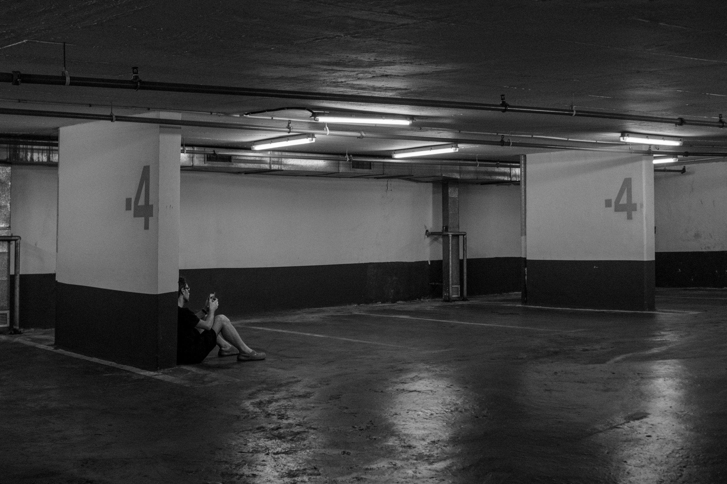 A person sits alone against a pillar in an empty underground parking garage marked level -4, lit by fluorescent lights