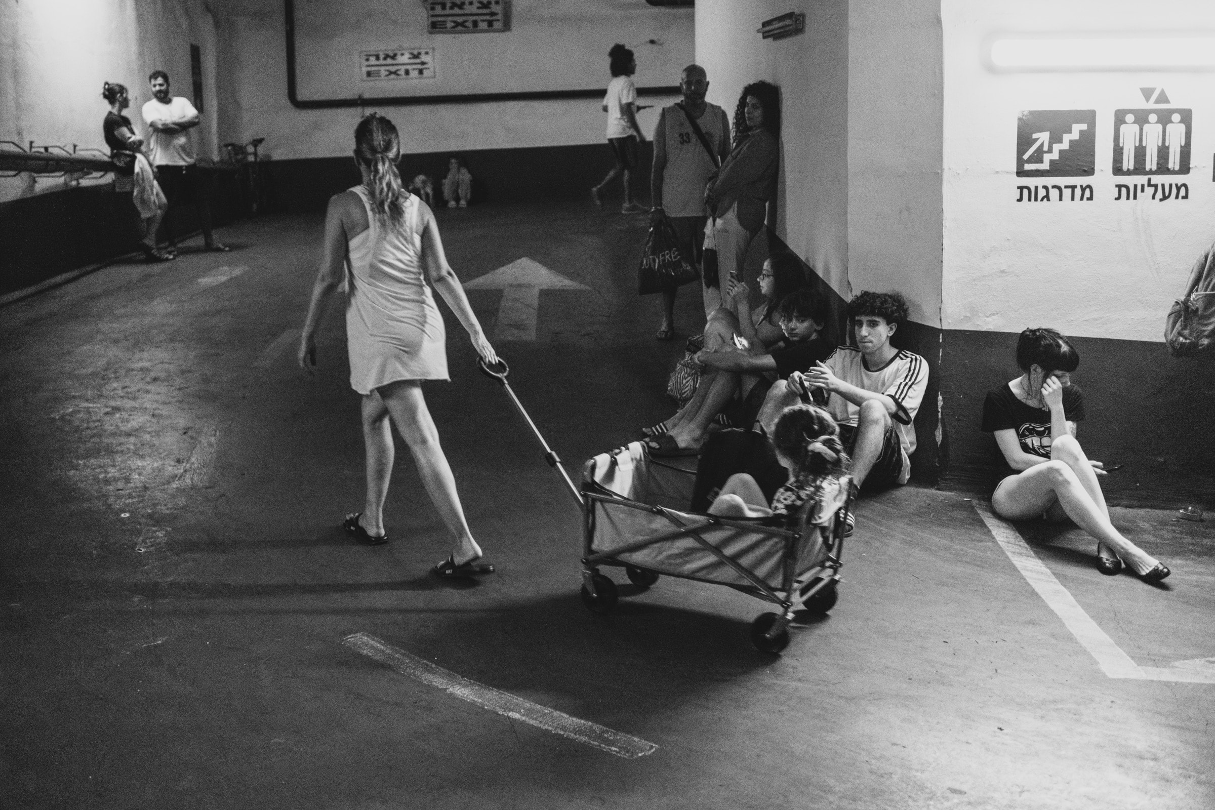 A woman pulling a wagon with a dog walks through a parking garage as a group of teenagers sits by an exit sign