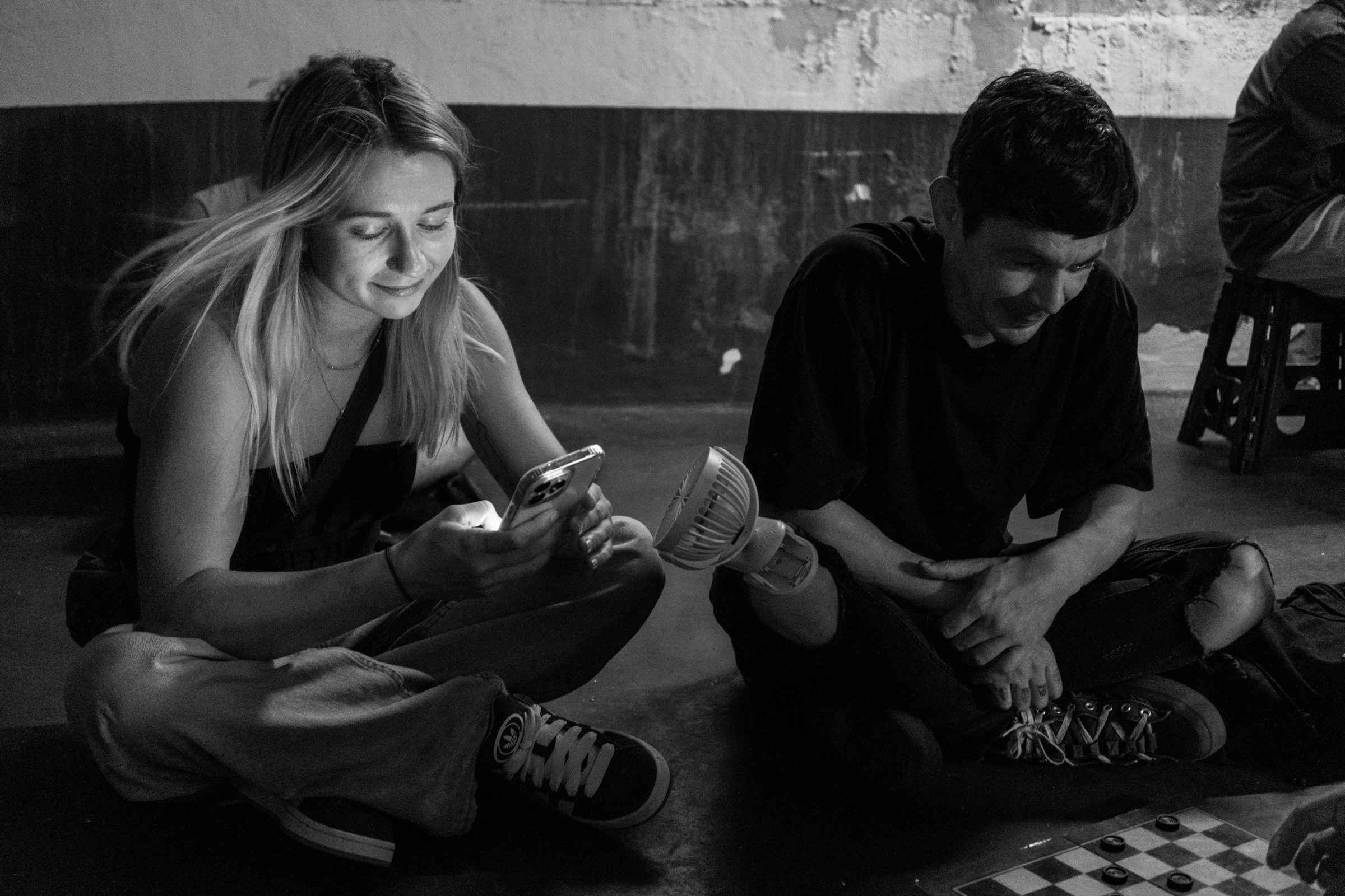 Two teenagers sit cross-legged on the floor of a parking garage, the girl smiling at her phone