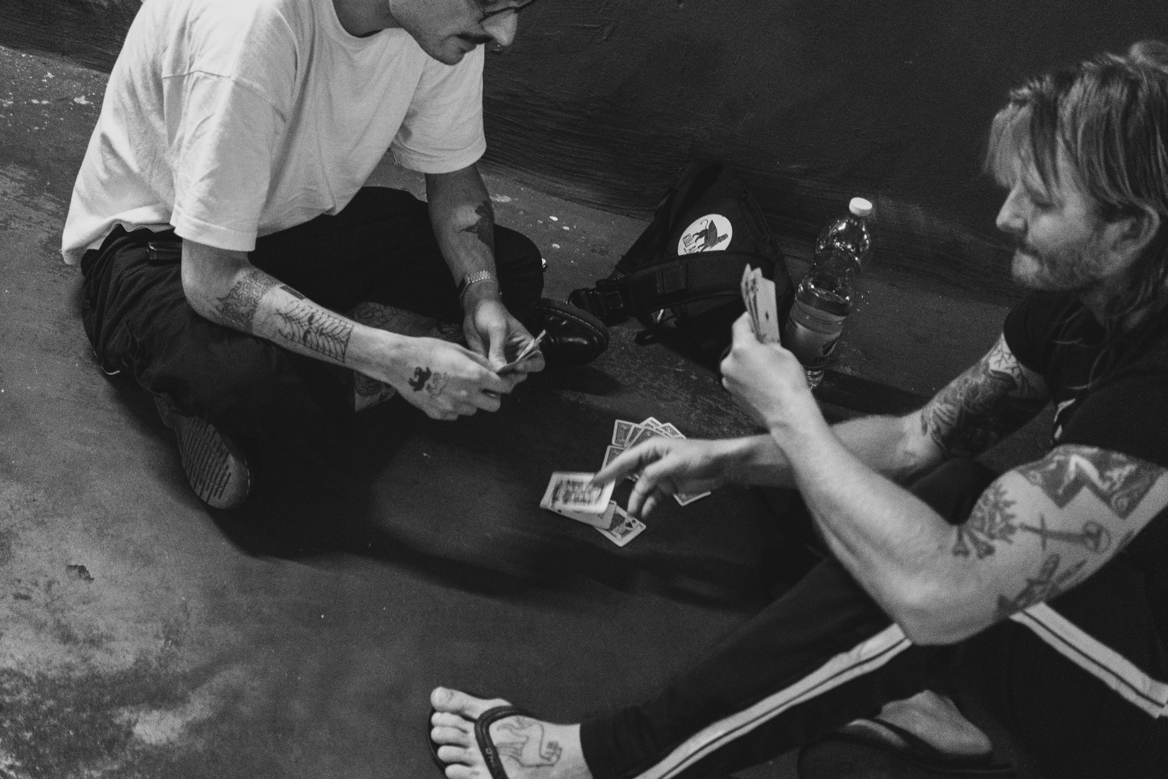 Two tattooed men sit on the concrete floor of a parking garage playing cards