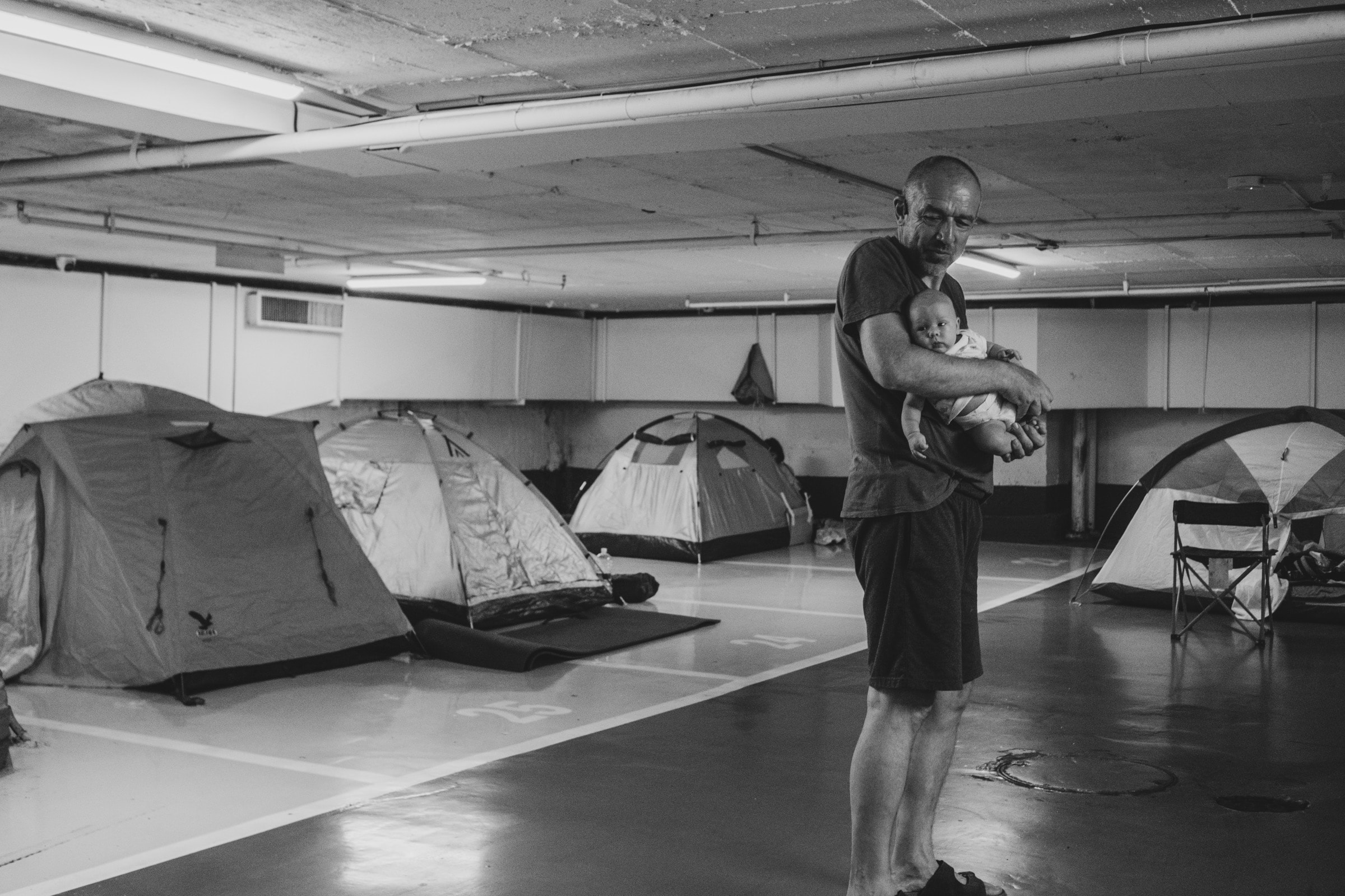 A man holds a baby while standing among rows of tents set up in a parking garage shelter