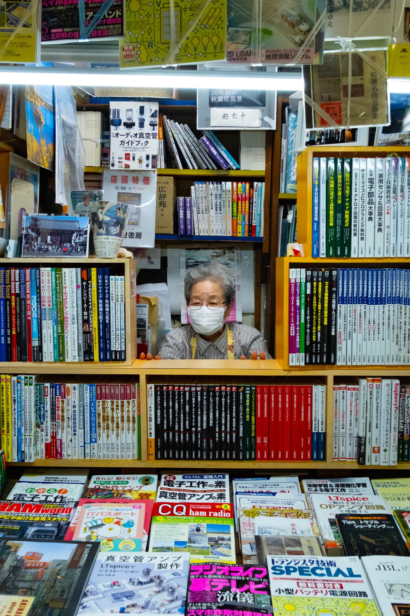 An elderly woman in a mask peers out from a small gap between shelves crammed with books and electronics magazines