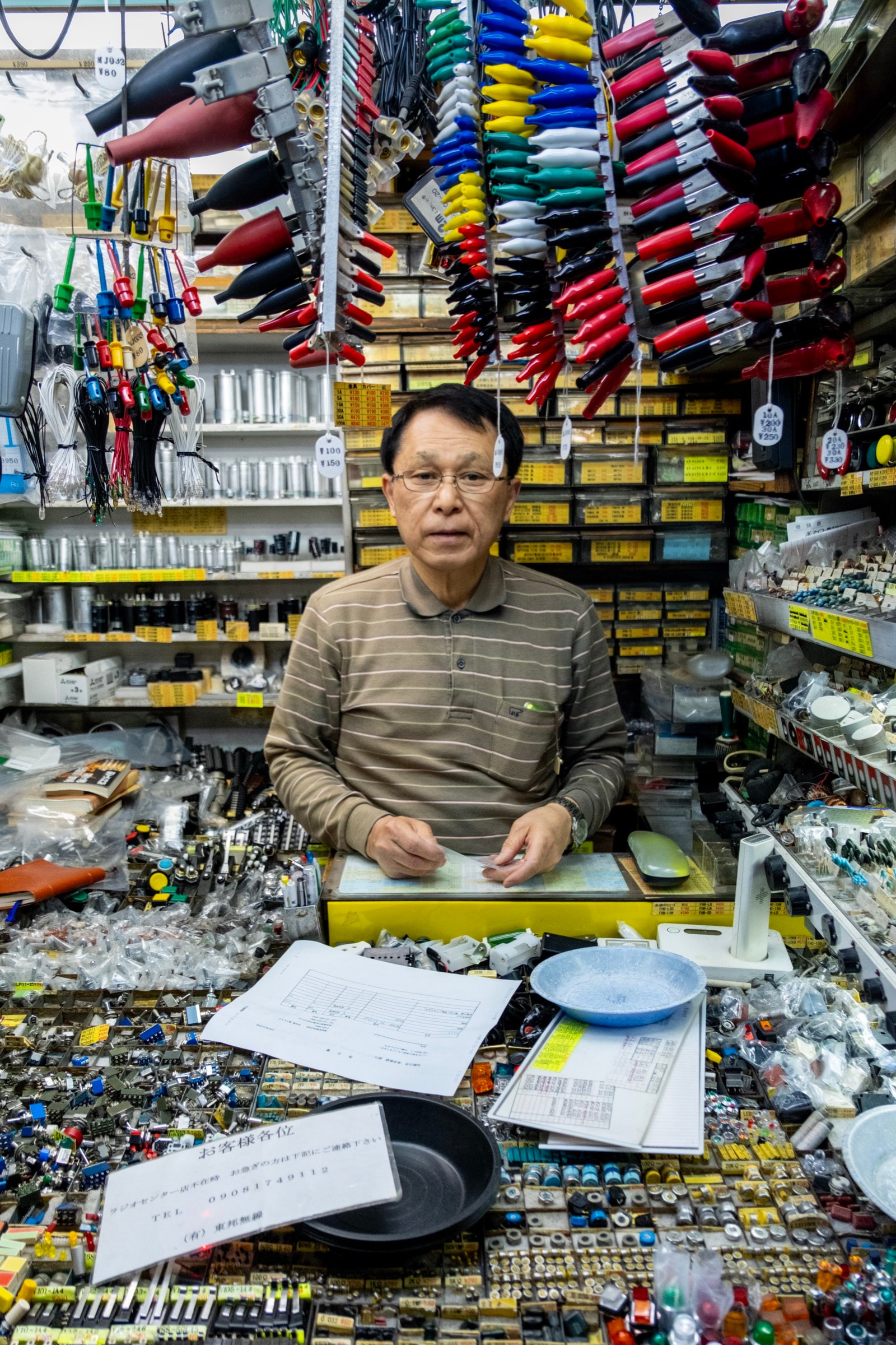 A man in glasses sits surrounded by thousands of tiny electronic parts, tools hanging from the ceiling in a packed Akihabara shop