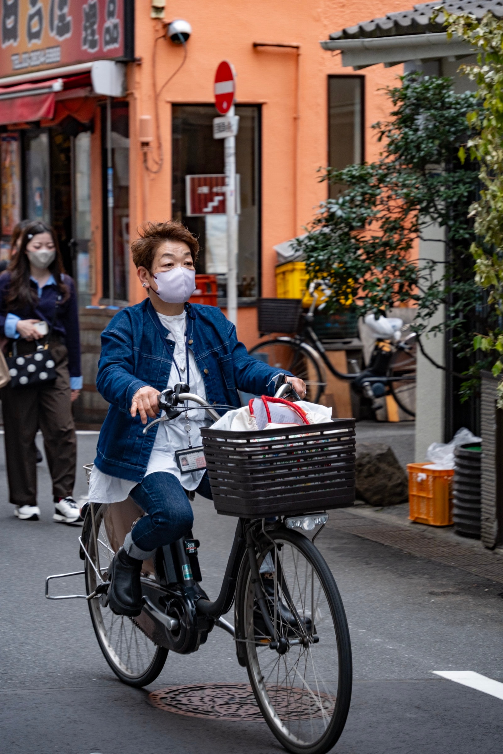 A woman in a denim jacket rides a bicycle with a basket down a narrow street past an orange building