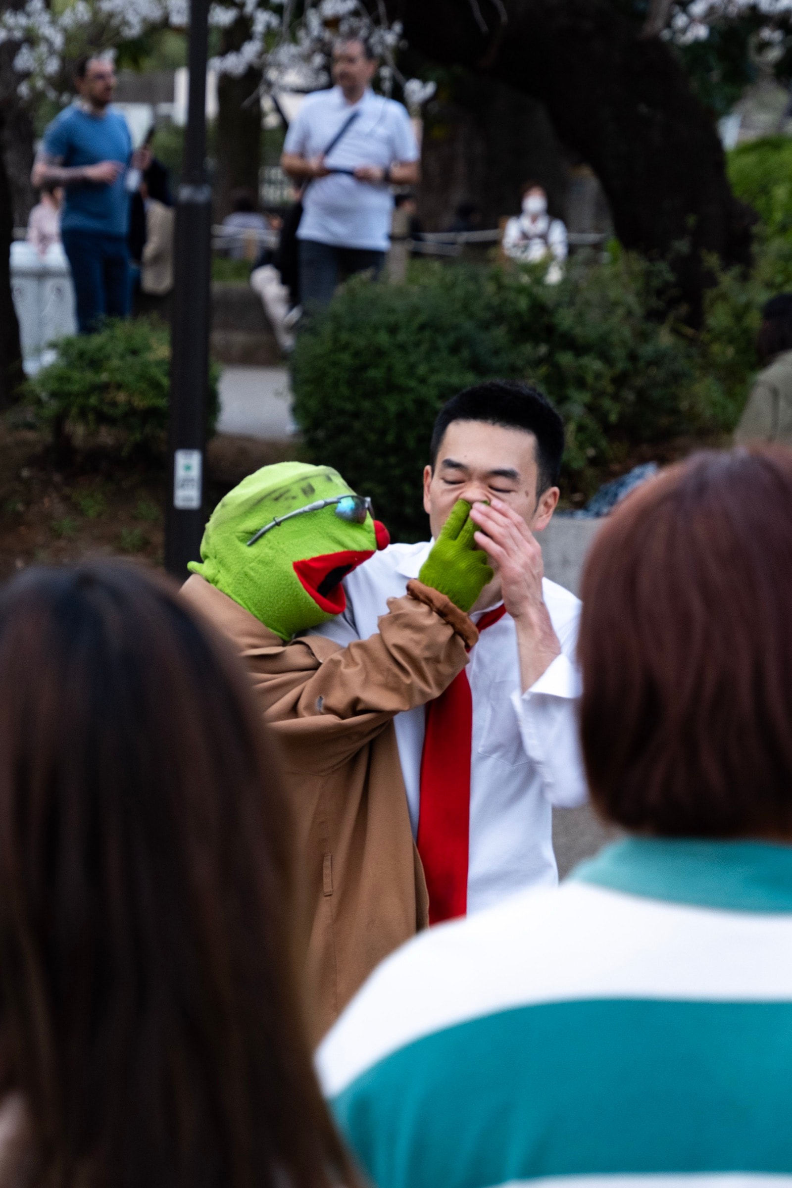 A street performer with a Kermit-the-Frog puppet pinches its green hand against his nose as a crowd watches in a park