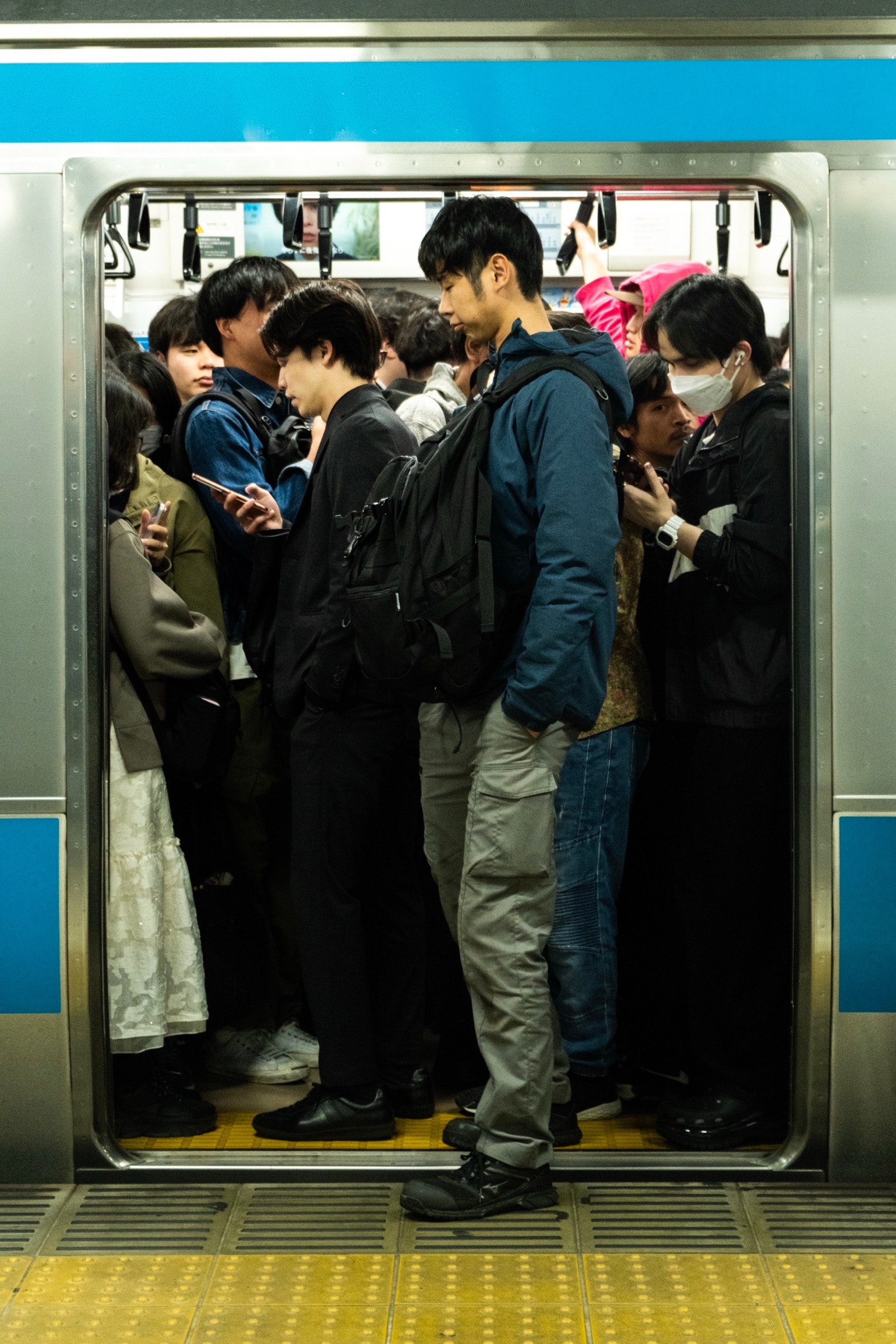 Commuters stand packed shoulder to shoulder in a train car with blue-striped doors, one man's foot on the yellow platform edge