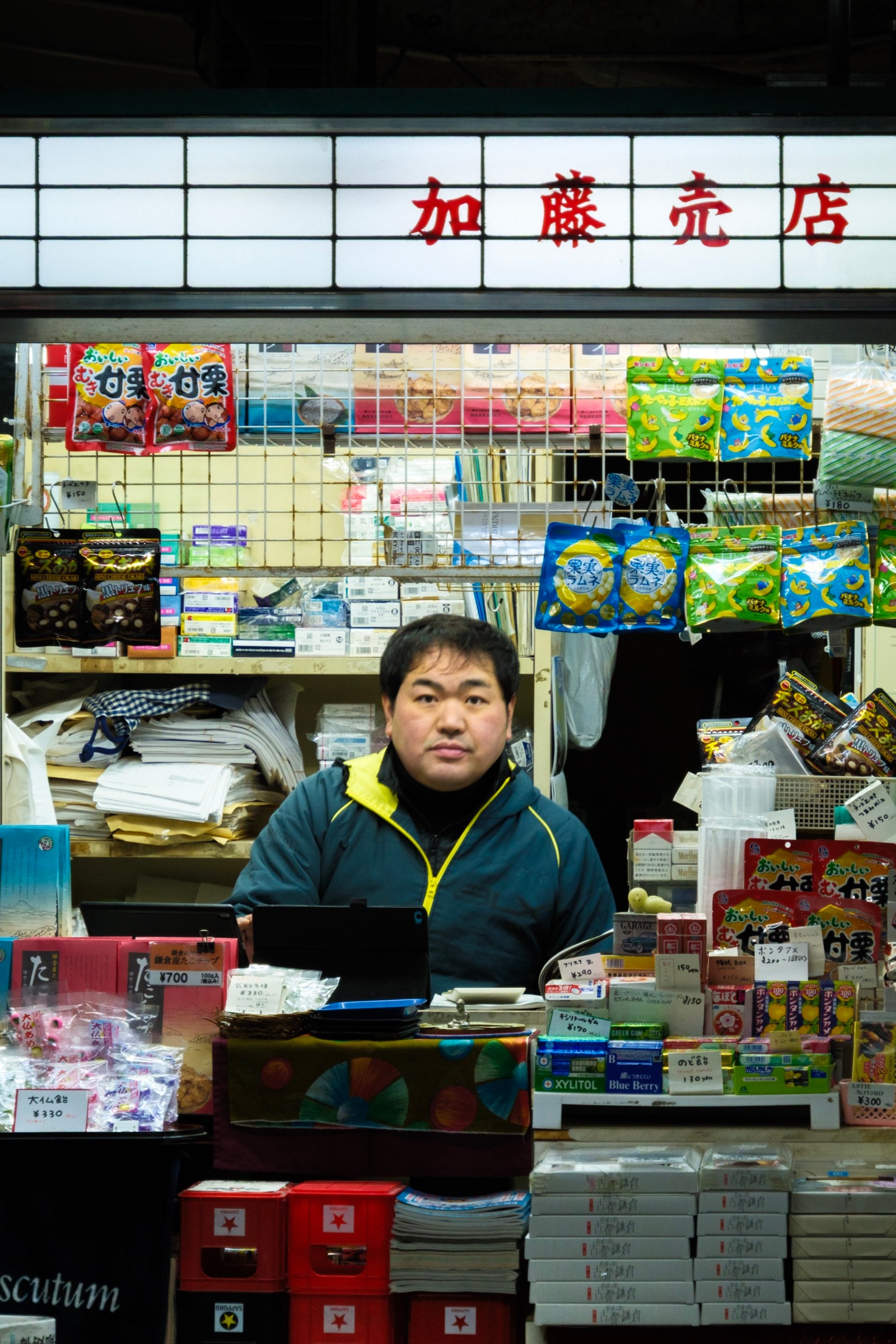 A vendor in a yellow-trimmed jacket stands behind the counter of a small station kiosk stocked with snacks and souvenirs