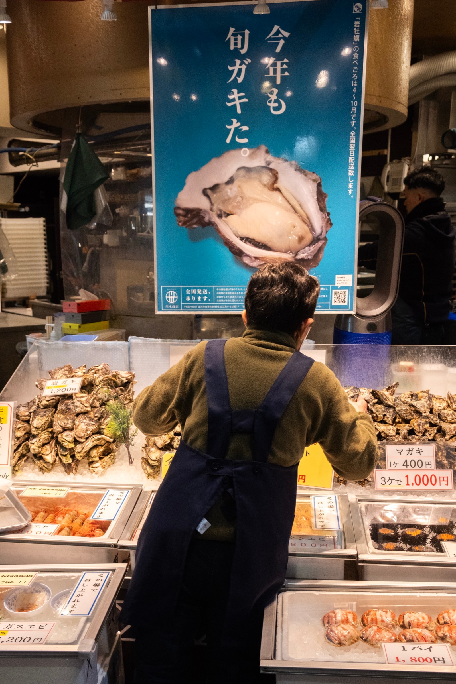 A vendor in an apron stands behind trays of oysters and seafood beneath a large blue poster advertising seasonal oysters
