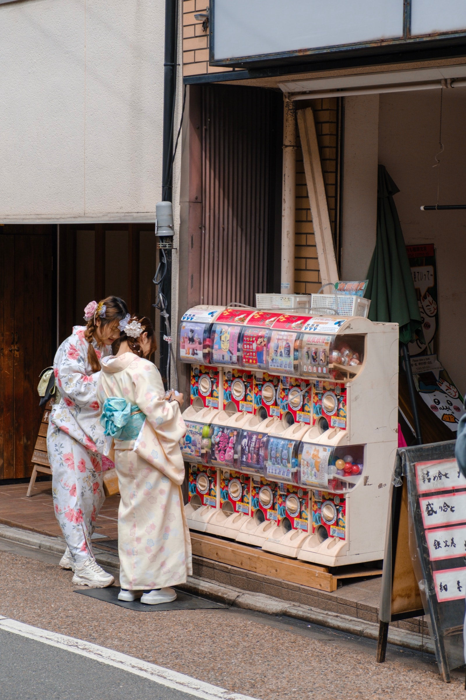Two young women in floral kimonos browse a row of gachapon capsule-toy machines on a street