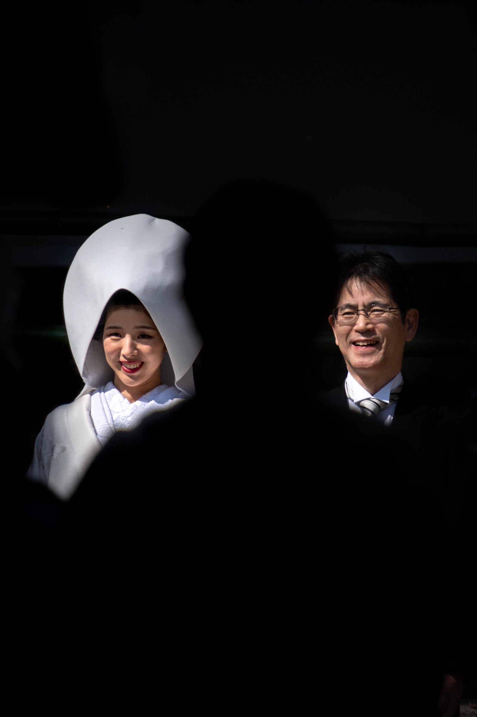 A bride in a watabōshi and a man in glasses laugh together, seen through a gap in a crowd of silhouettes