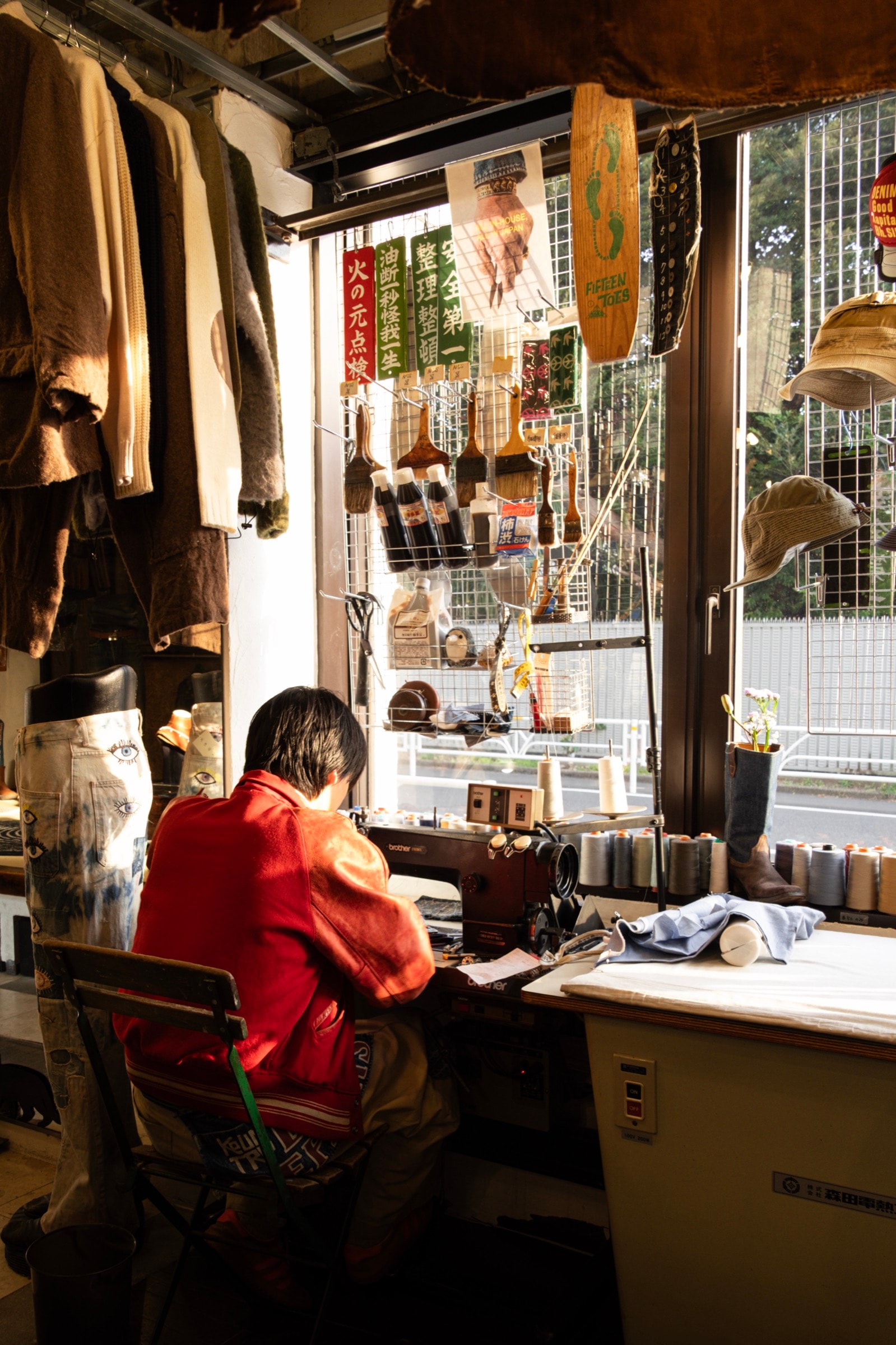 A woman in a red shirt sits at a sewing machine by a window in a cluttered tailor's workshop hung with clothes and hats