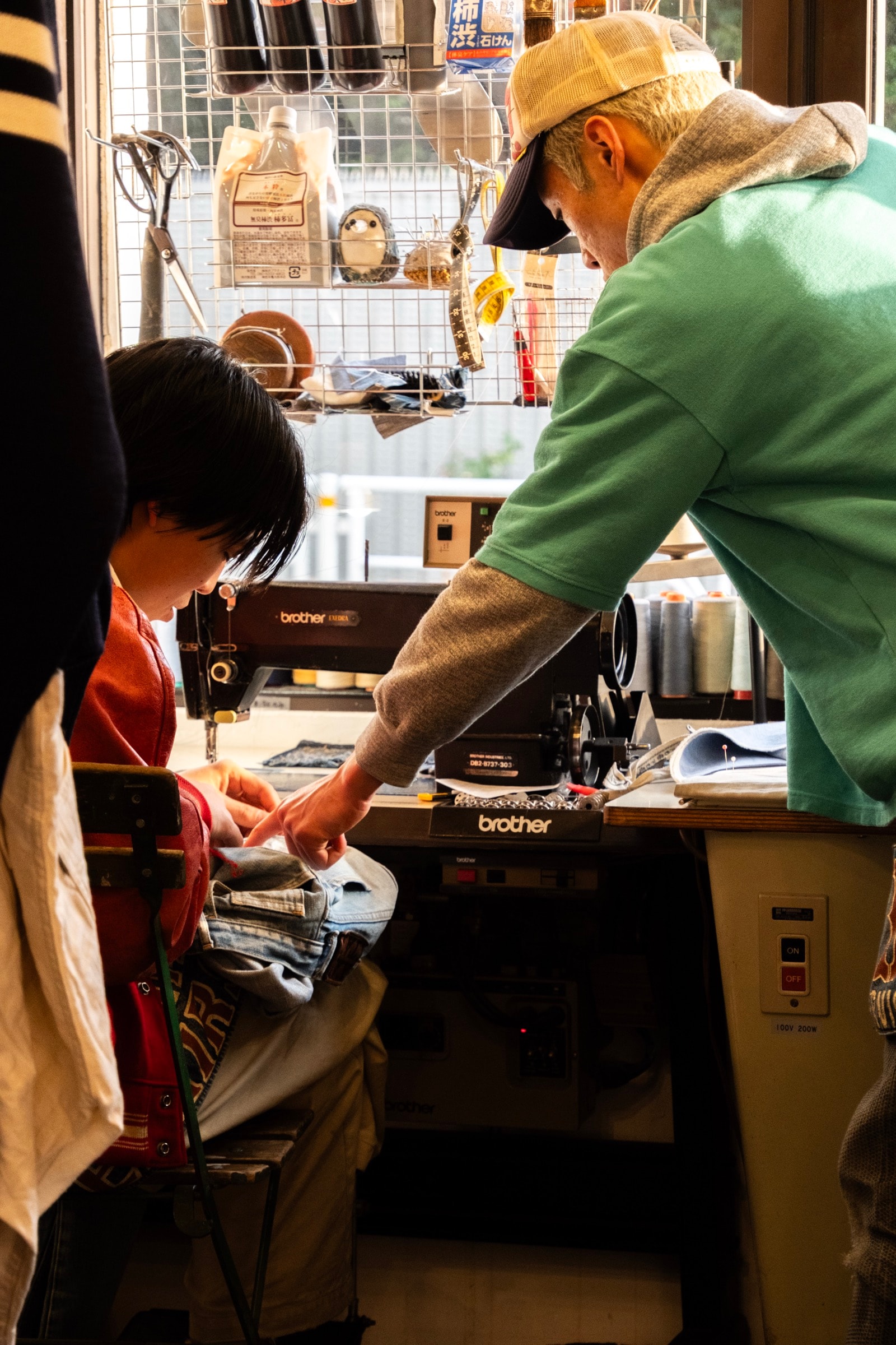 A man in a green sweatshirt and cap hands fabric to a woman seated at a sewing machine in a small workshop