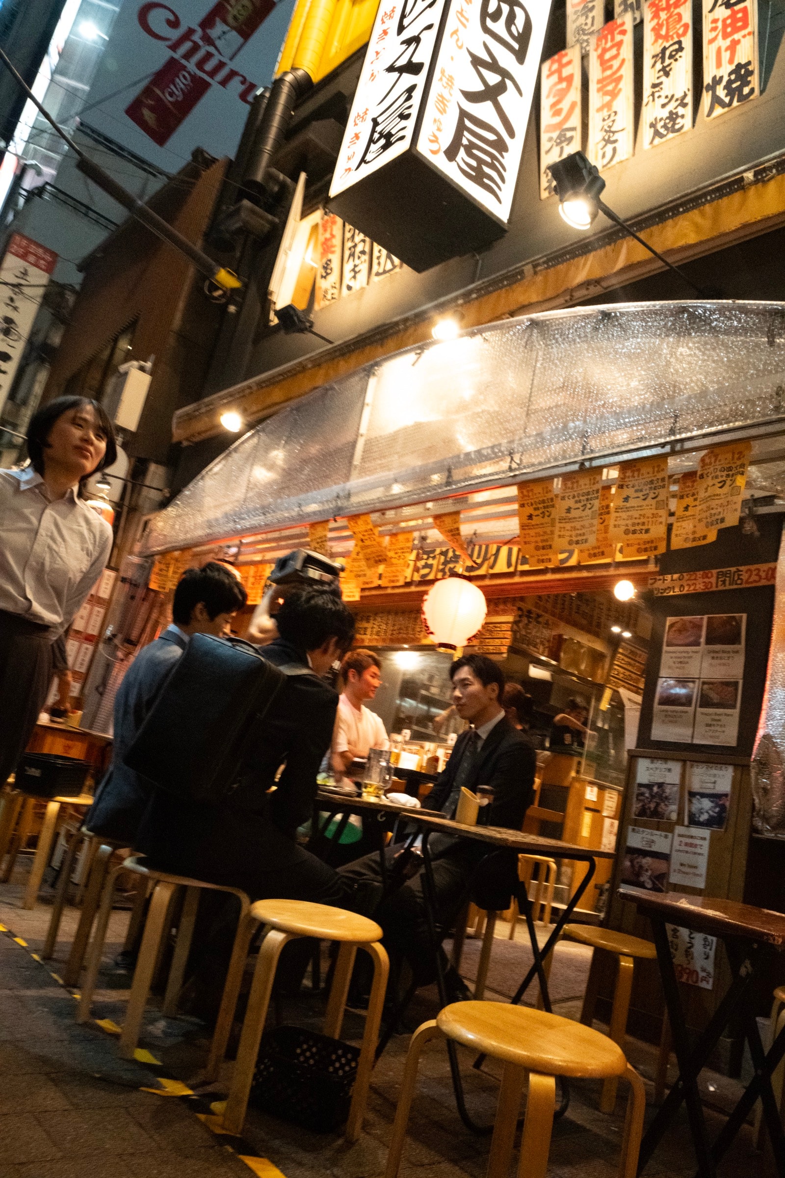 Salarymen drink and chat at a small open-front izakaya with wooden stools and Japanese signs stacked above