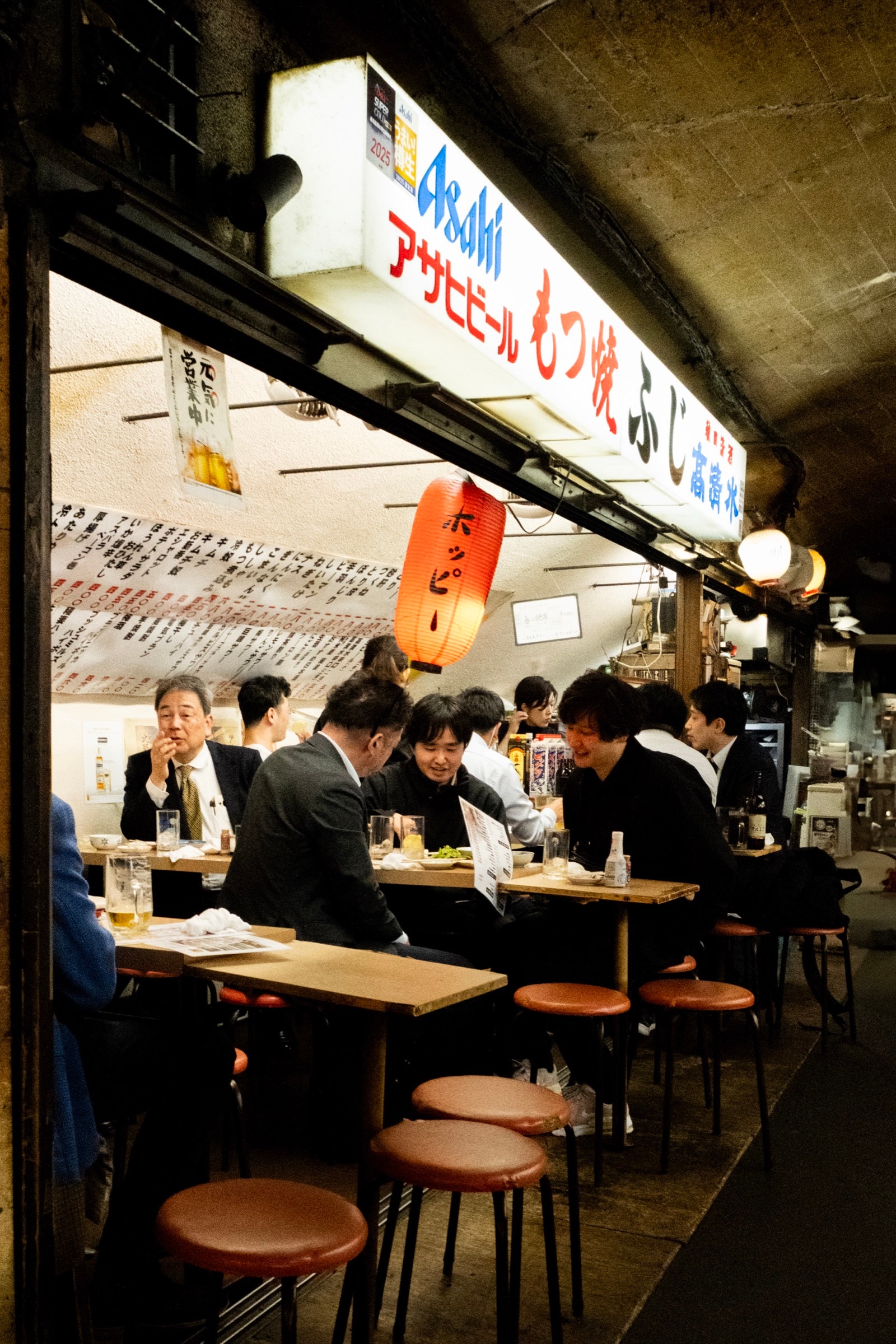 Office workers crowd a narrow motsuyaki grill bar under an Asahi beer sign and a glowing Hoppy lantern