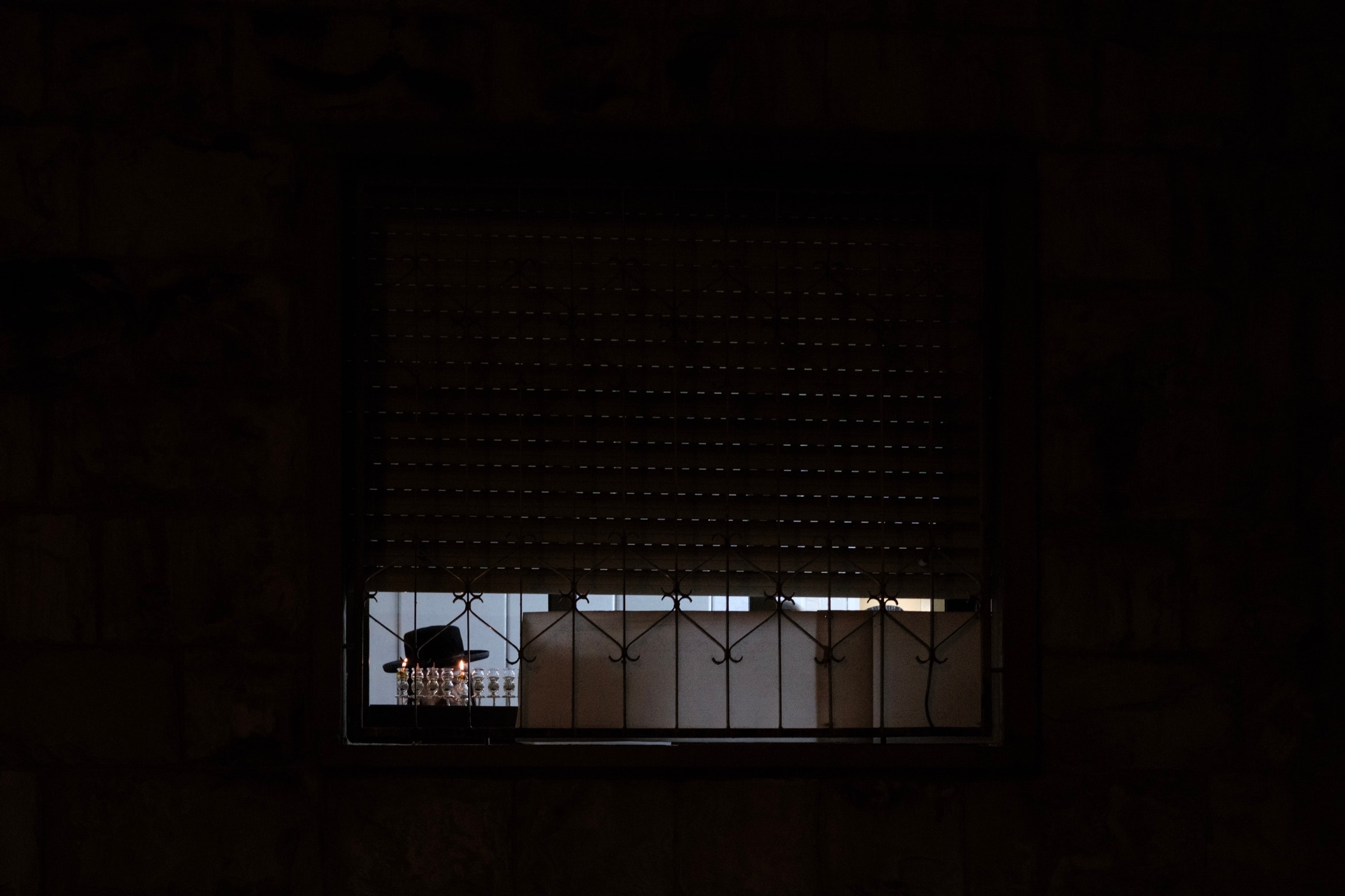 A lit menorah seen through a window with half-closed blinds and iron bars, the top of a black hat just visible below