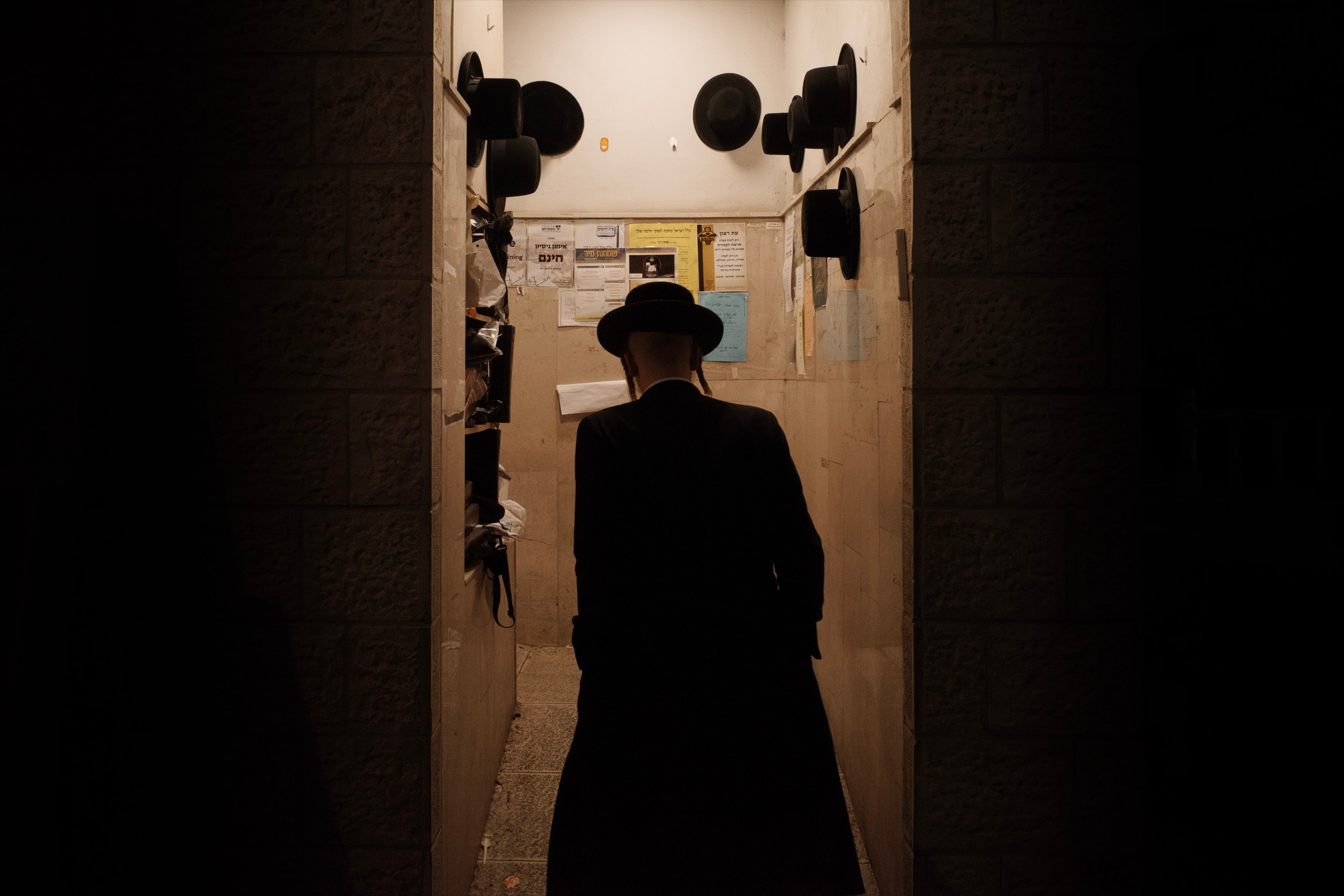 A man in a black hat walks into a narrow hallway entrance with black hats hanging on hooks along both walls