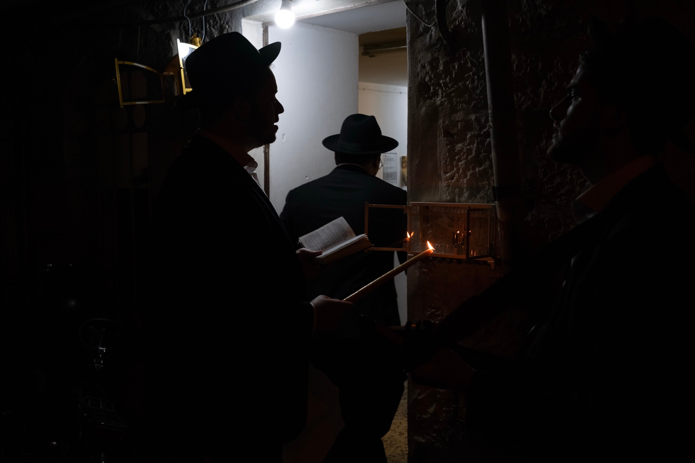 Two silhouetted men in hats, one holding a candle and prayer book, light a menorah in a glass case on a narrow stone alleyway
