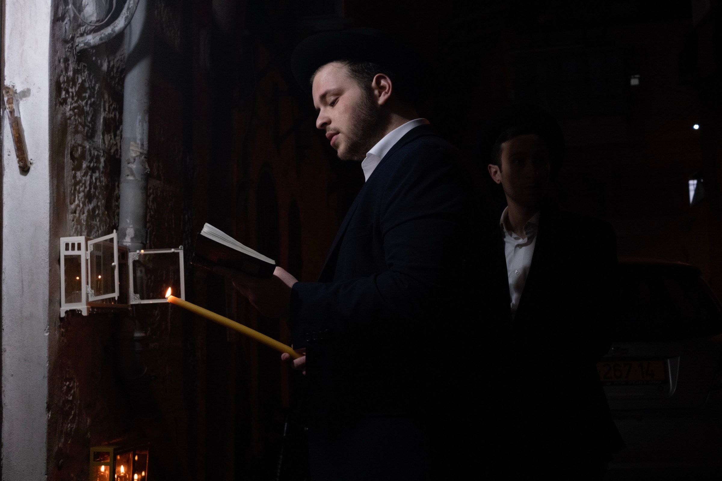 A young man reads from a prayer book by the light of a yellow candle beside a small glass-enclosed menorah mounted on a stone wall