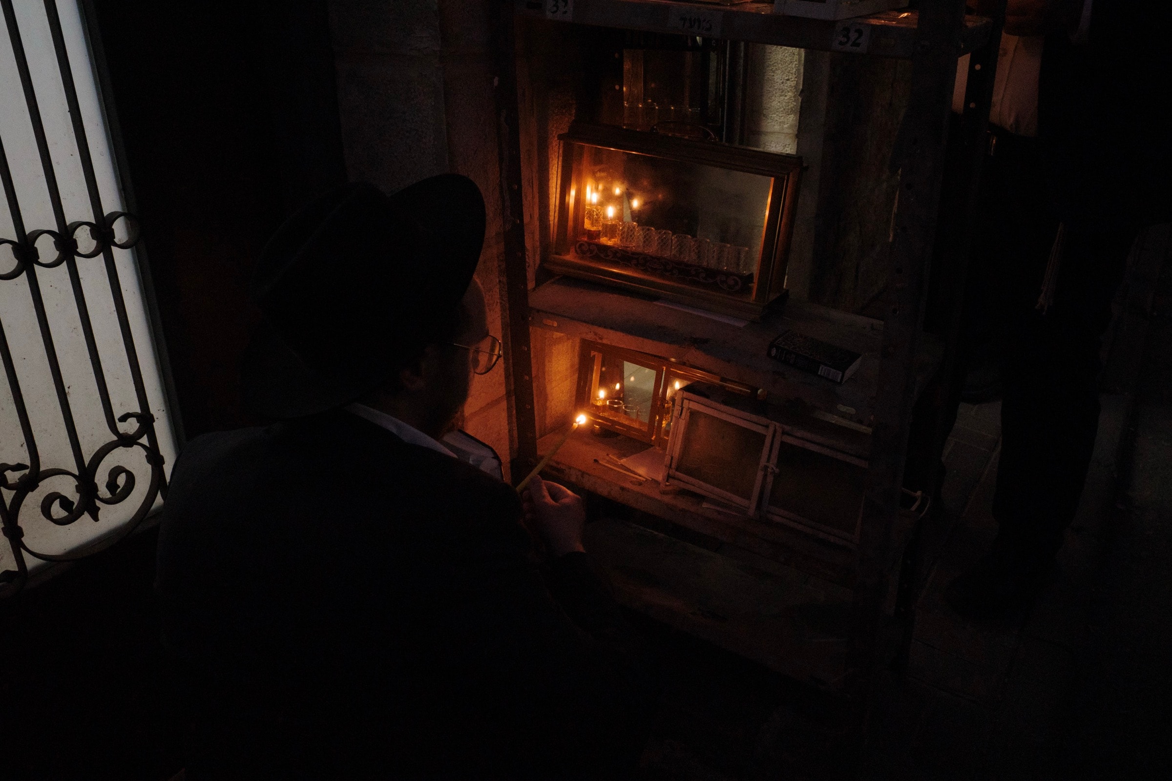 A man in a black hat crouches to light Hanukkah candles in glass-enclosed menorahs outside a doorway