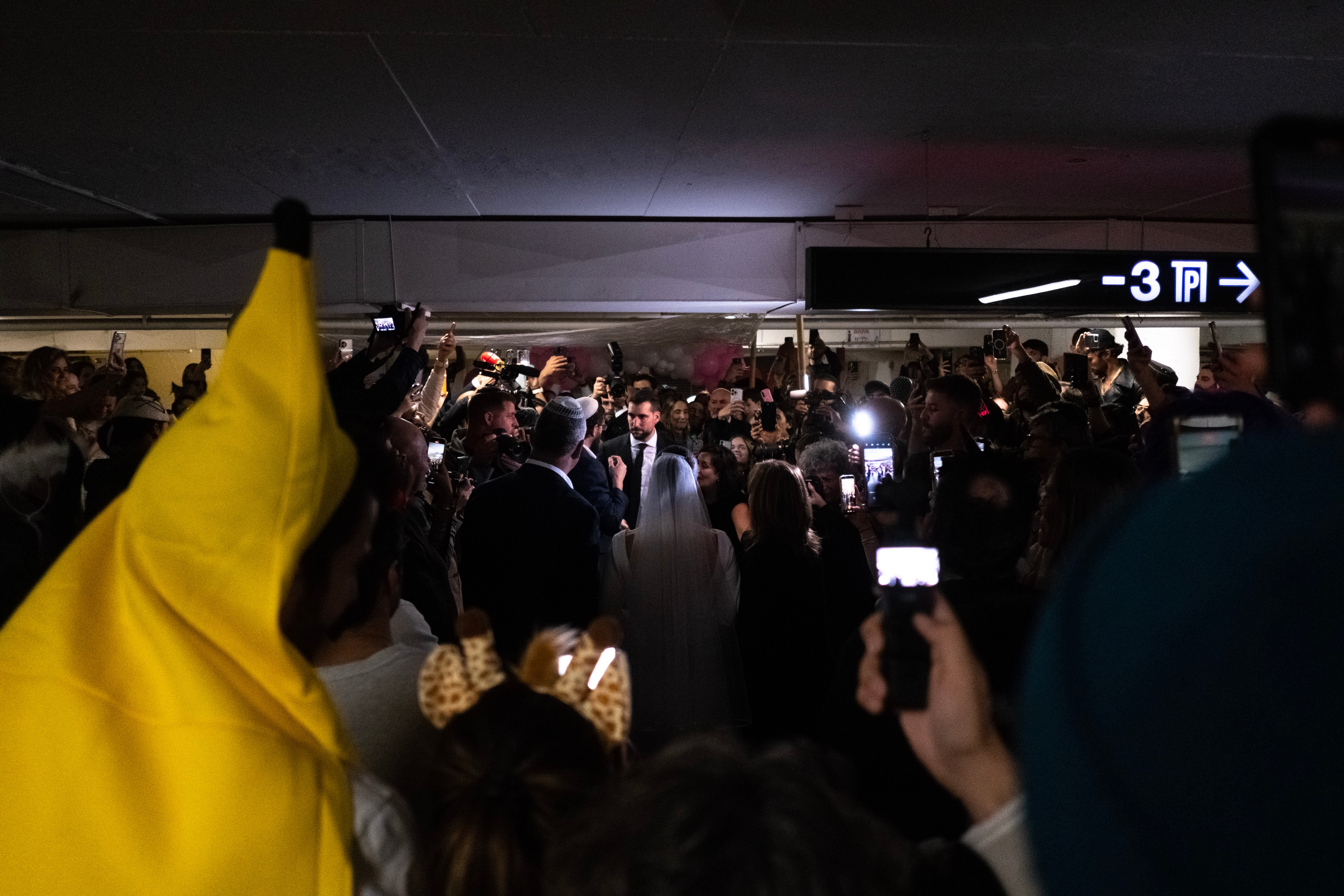 A crowd surrounds the couple in a dark parking garage, phones raised and flashing, a yellow flag waving in the foreground