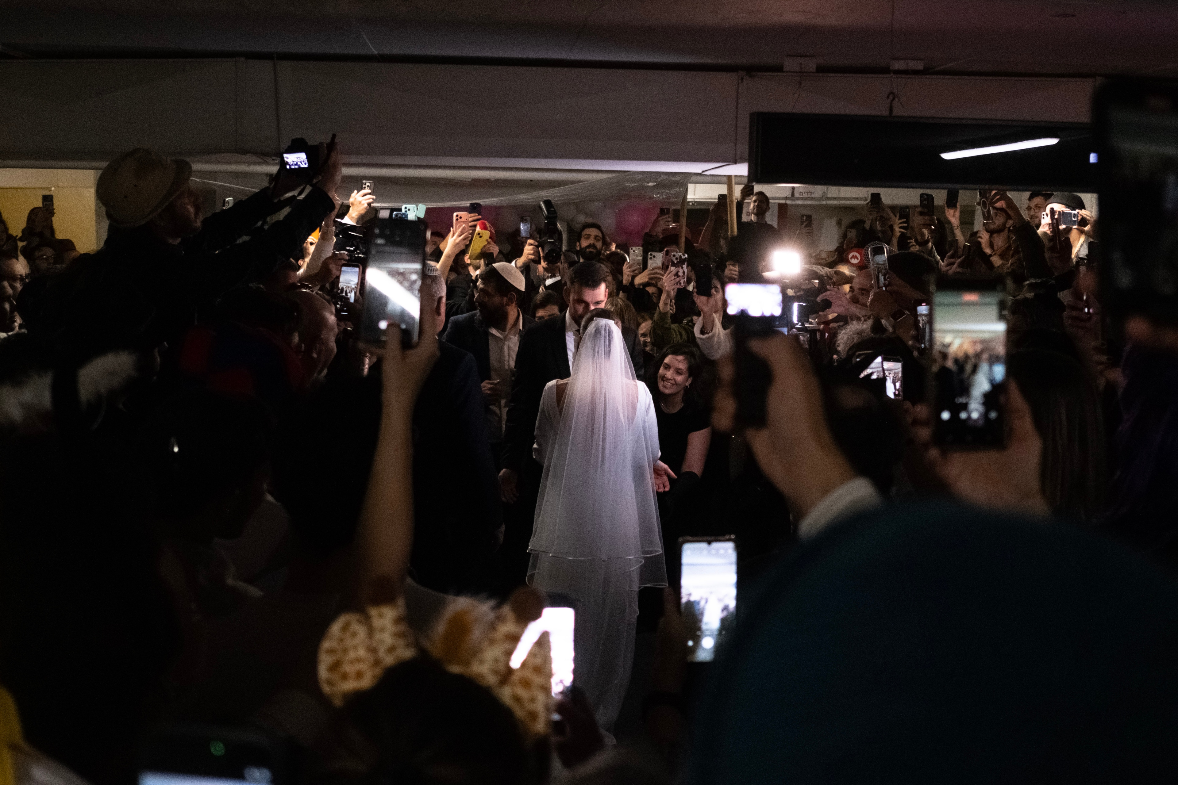 The bride in a long veil seen from behind, walking through a dense crowd of guests holding up phones in the parking garage