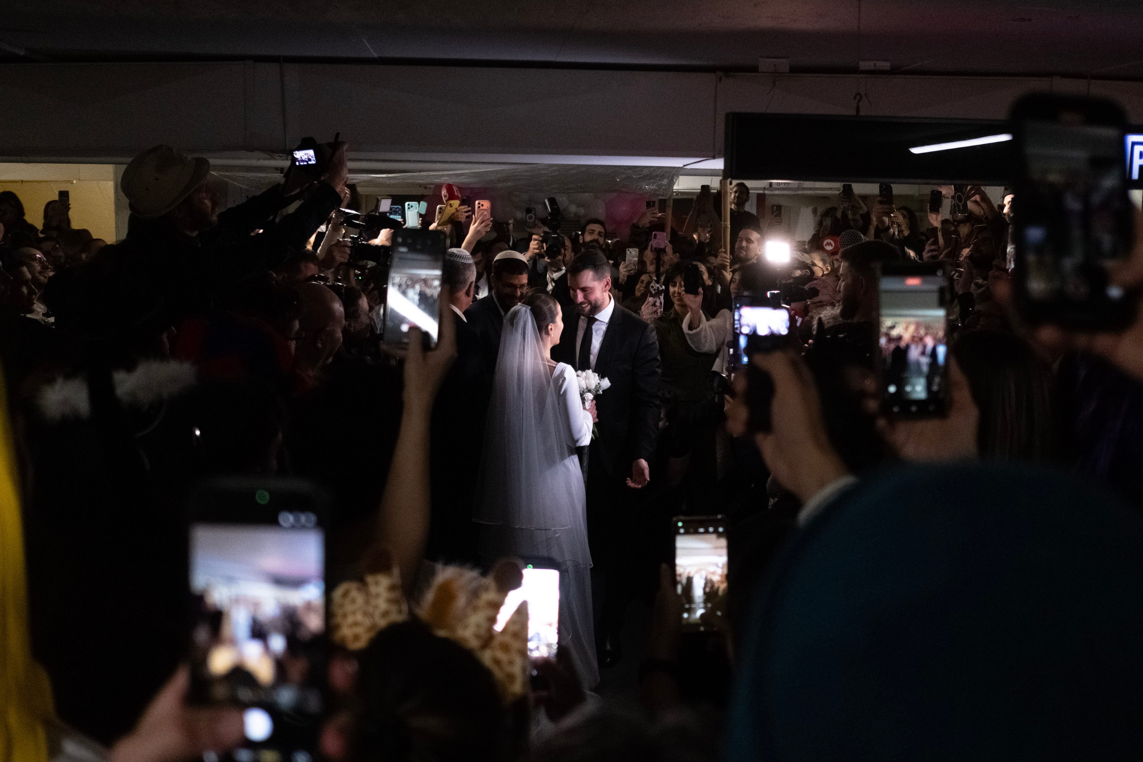 The couple face each other surrounded by guests recording on their phones, lit by a flash in the dim garage