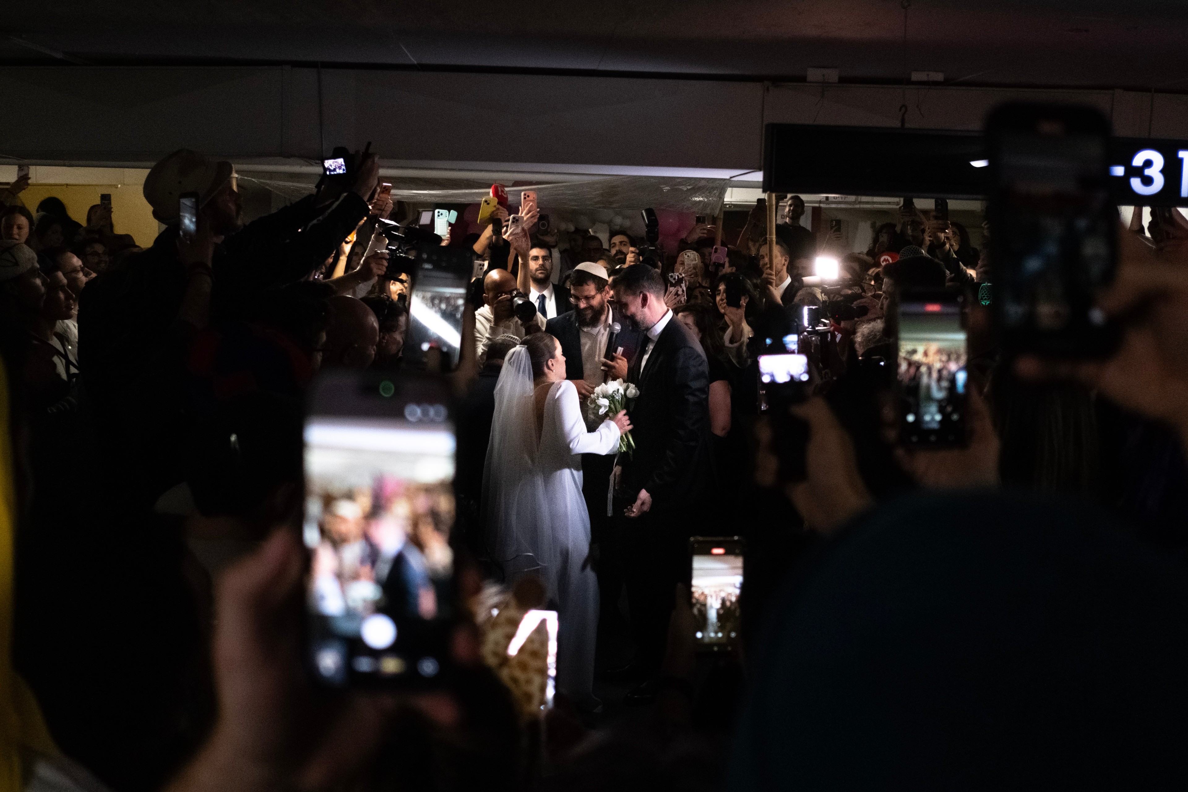 The bride and groom stand close together framed by a sea of raised phones and onlookers in the underground garage