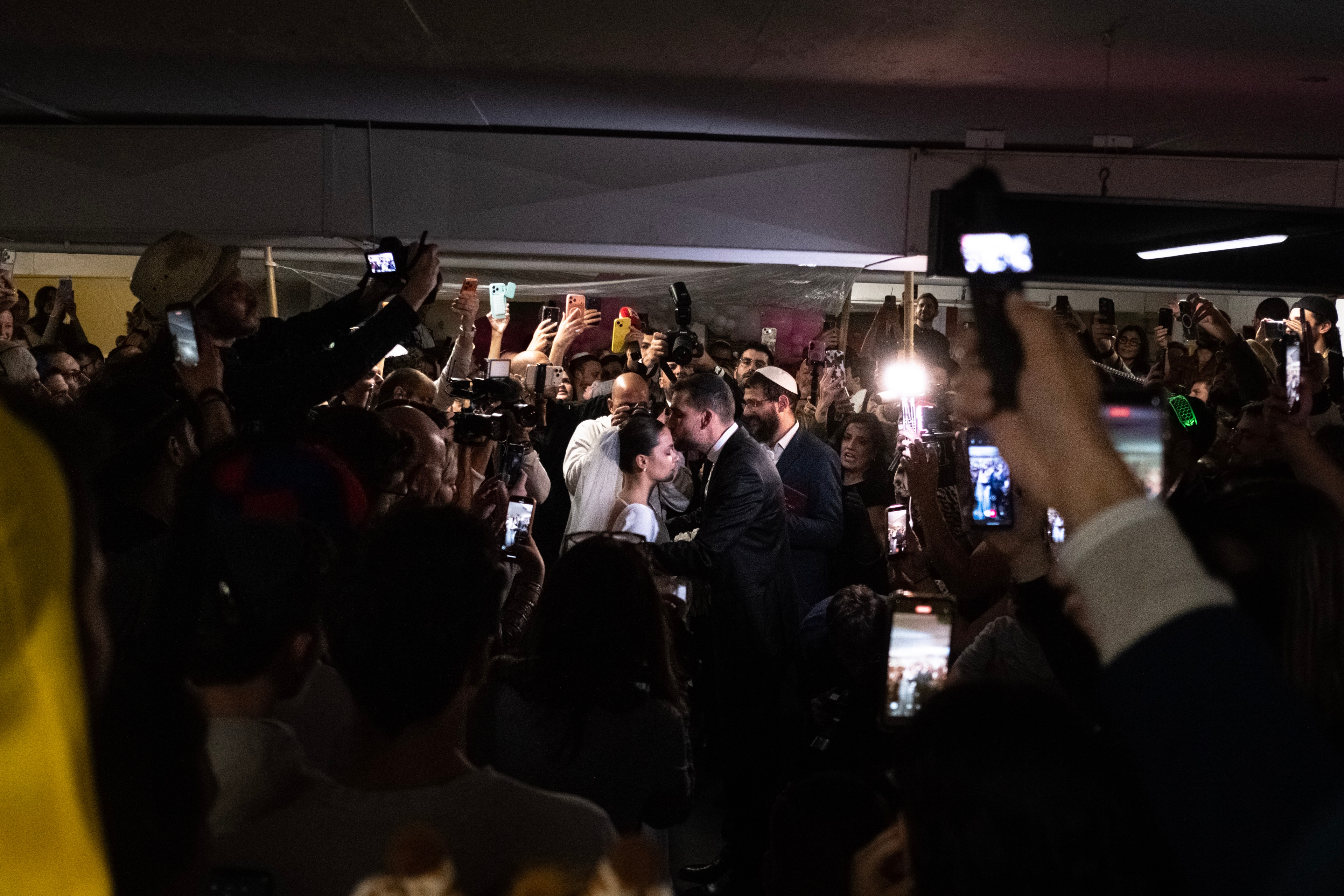 The couple kiss amid a cheering crowd in the parking garage, camera flashes and phone screens lighting the scene