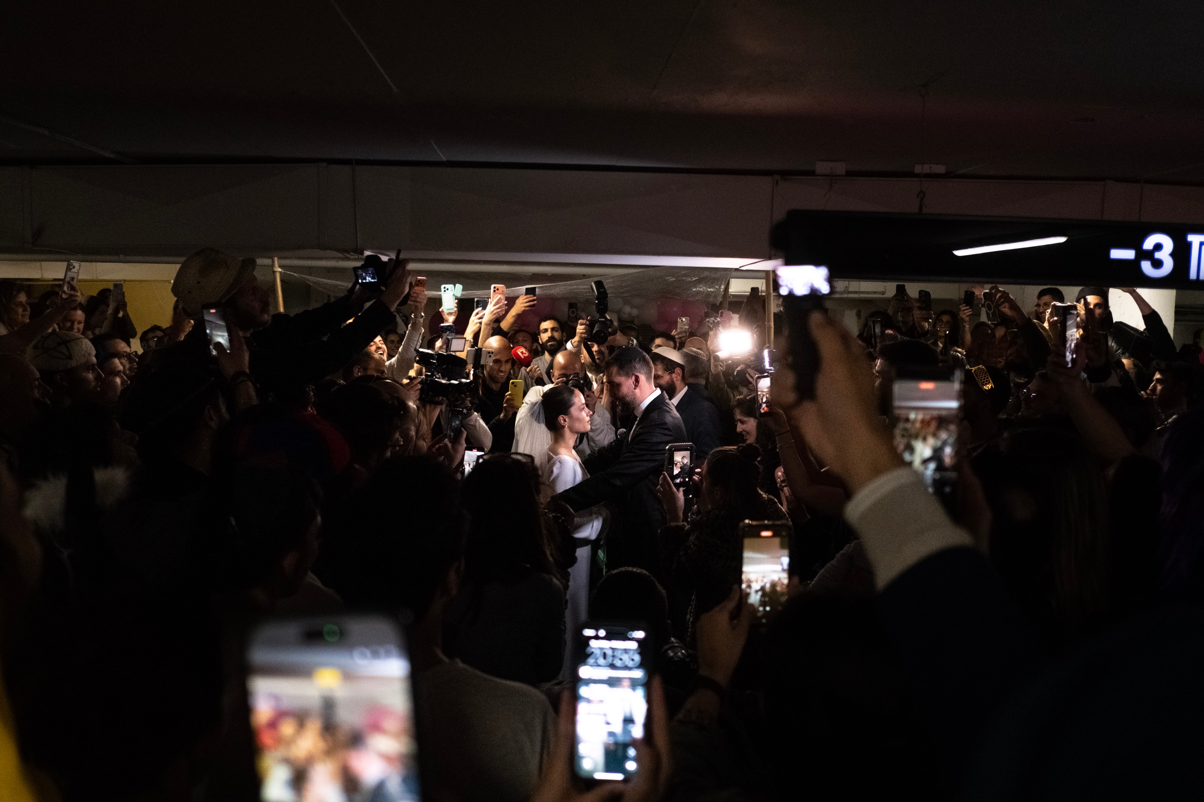 A wide shot of the couple embracing in the center of the crowd, arms and phones raised around them in the dark garage