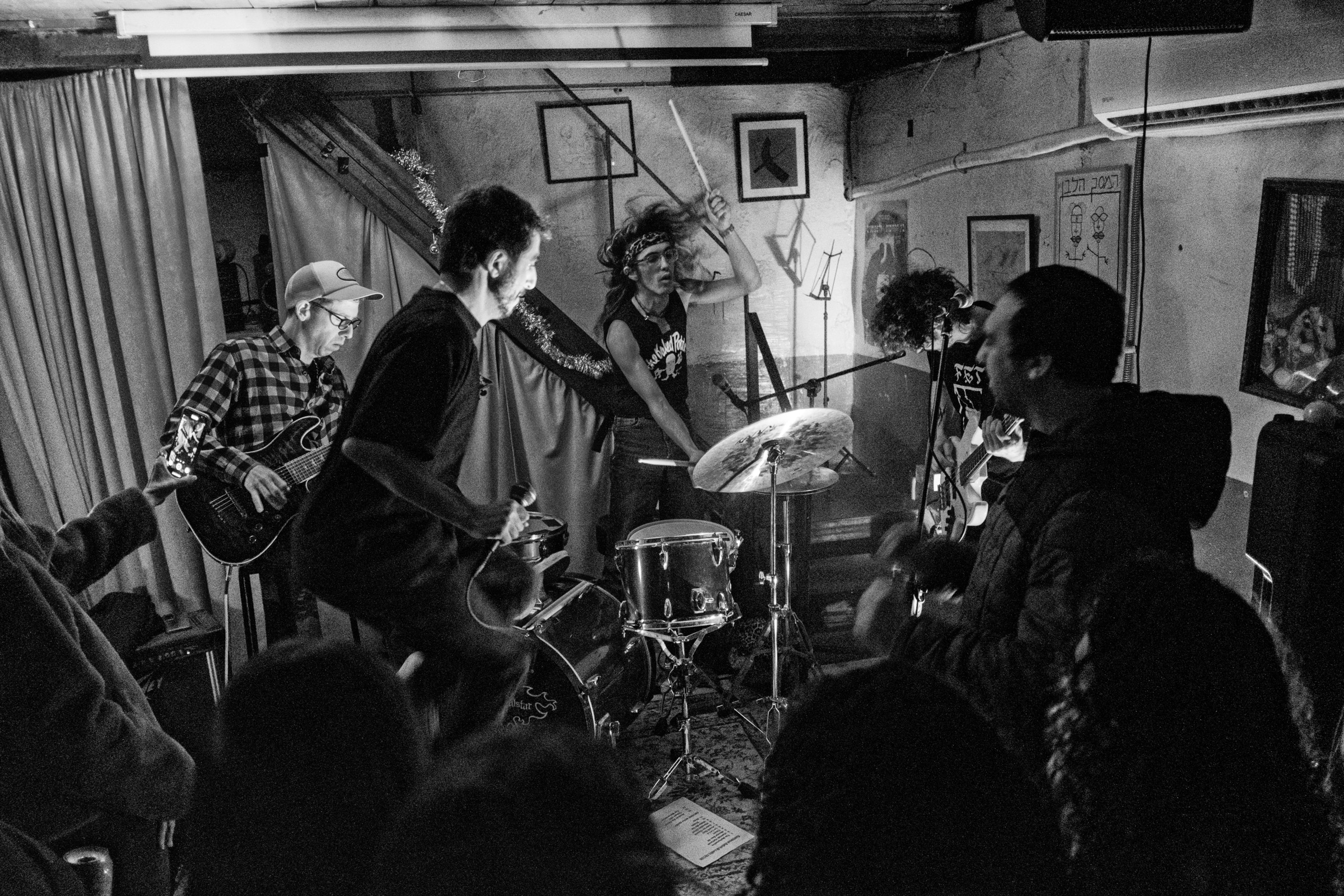 Band members face each other mid-performance in a basement, a woman on drums in the center, audience watching closely
