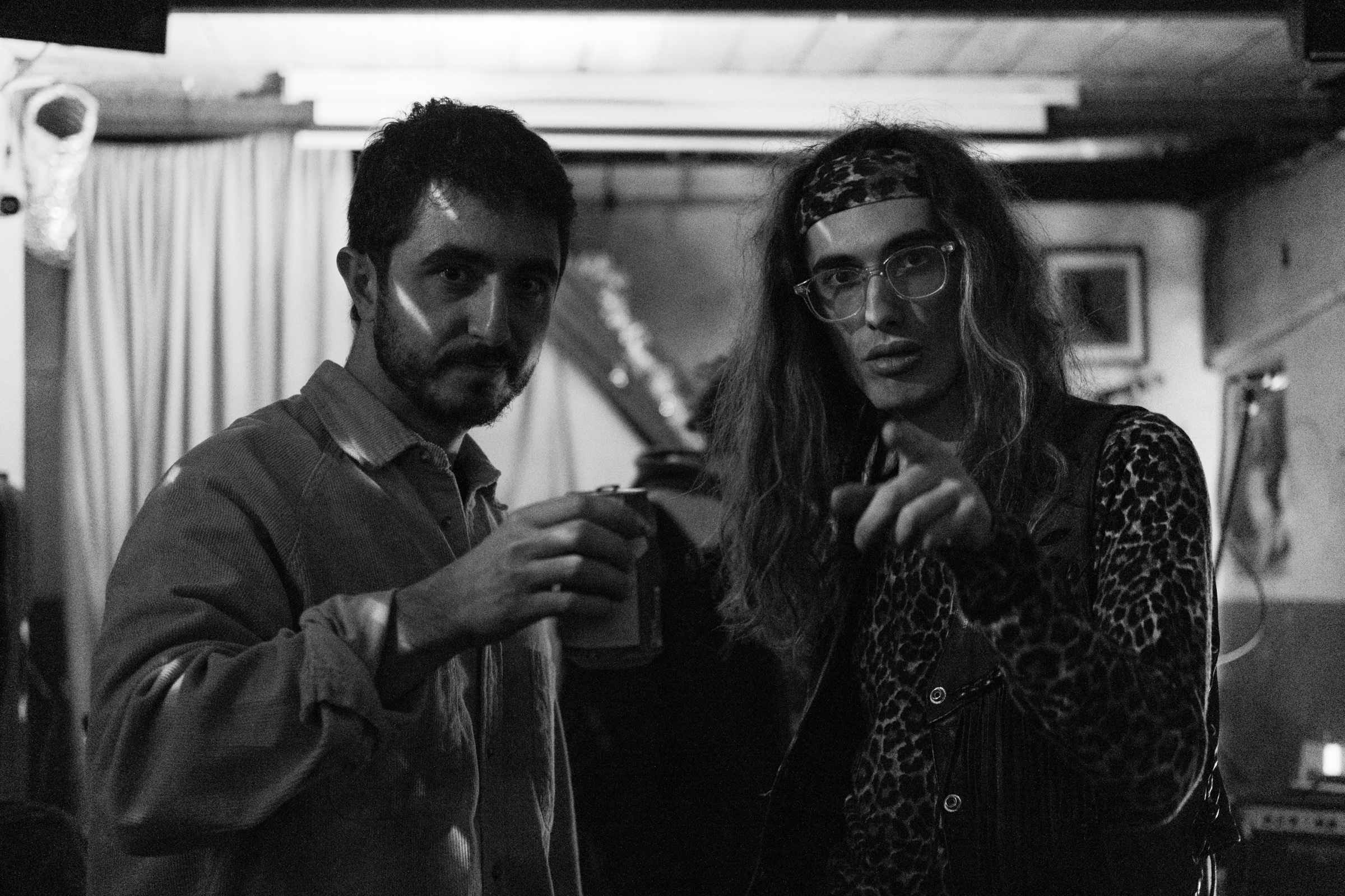 Two men pose backstage, one in a denim jacket holding a drink, the other in a leopard-print shirt and headband