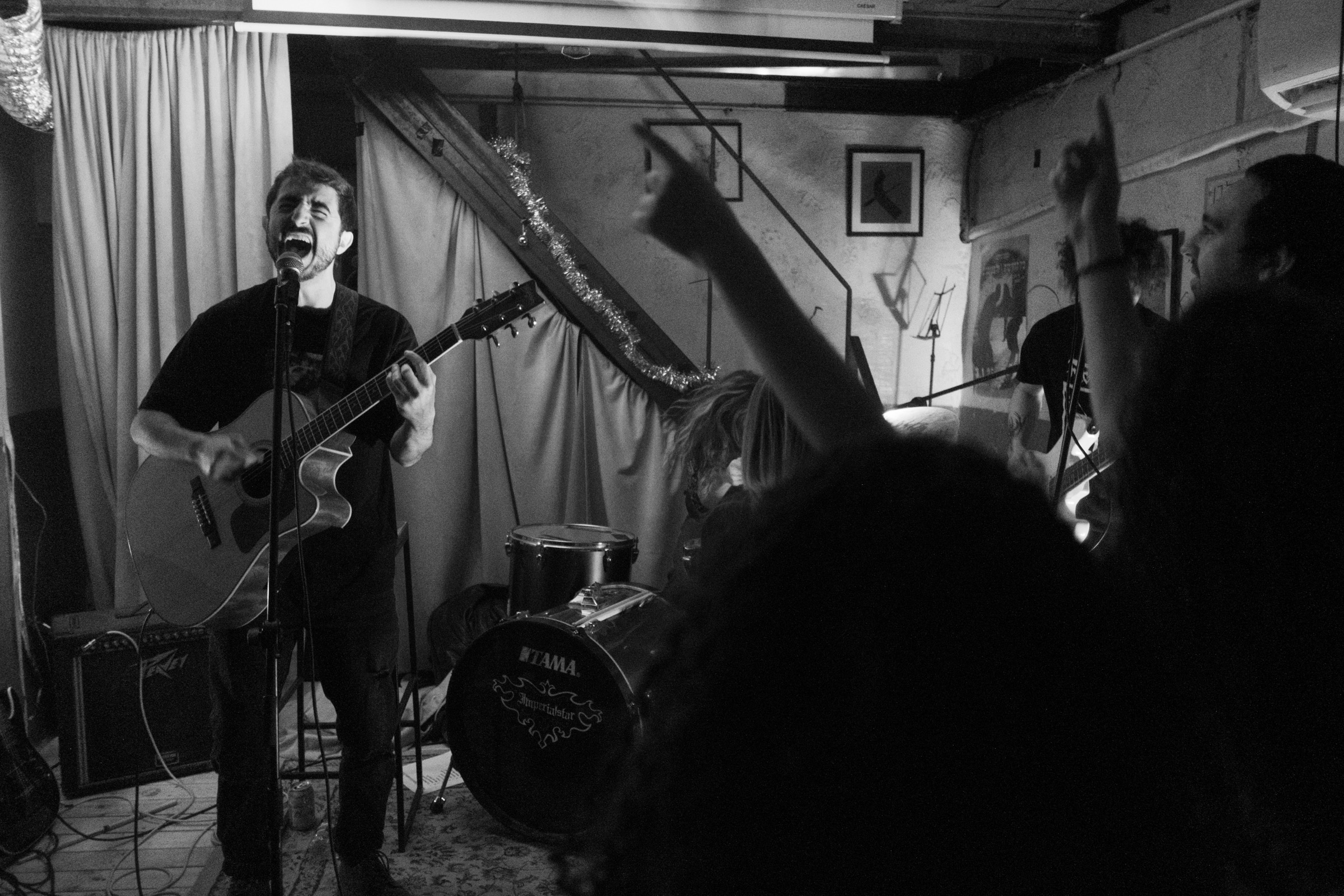 A singer screams into the mic while playing acoustic guitar on a small stage, audience hands raised