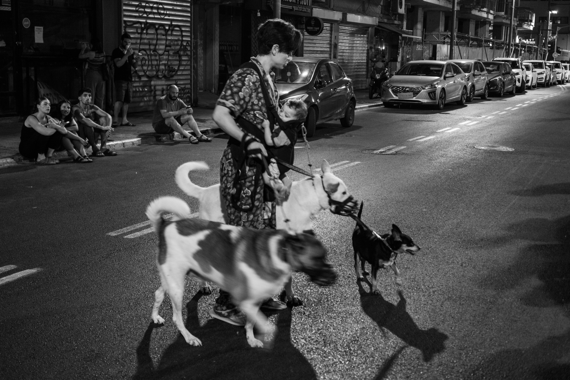A woman walks three dogs across a street at night while people sit on the sidewalk behind her