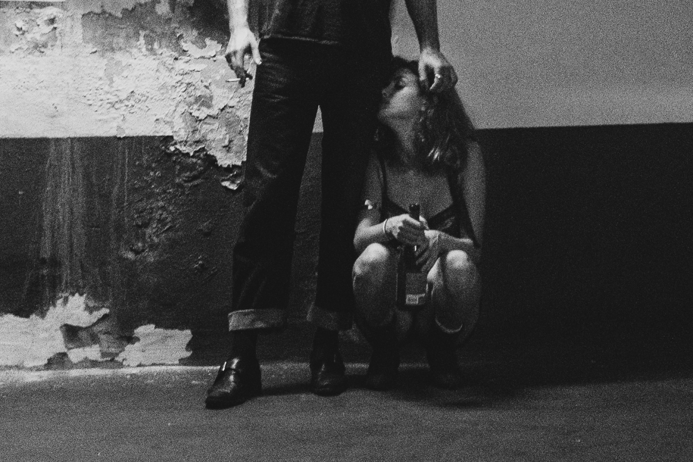 A person stands while a woman crouches against a peeling wall in a dimly lit parking garage, both holding cigarettes