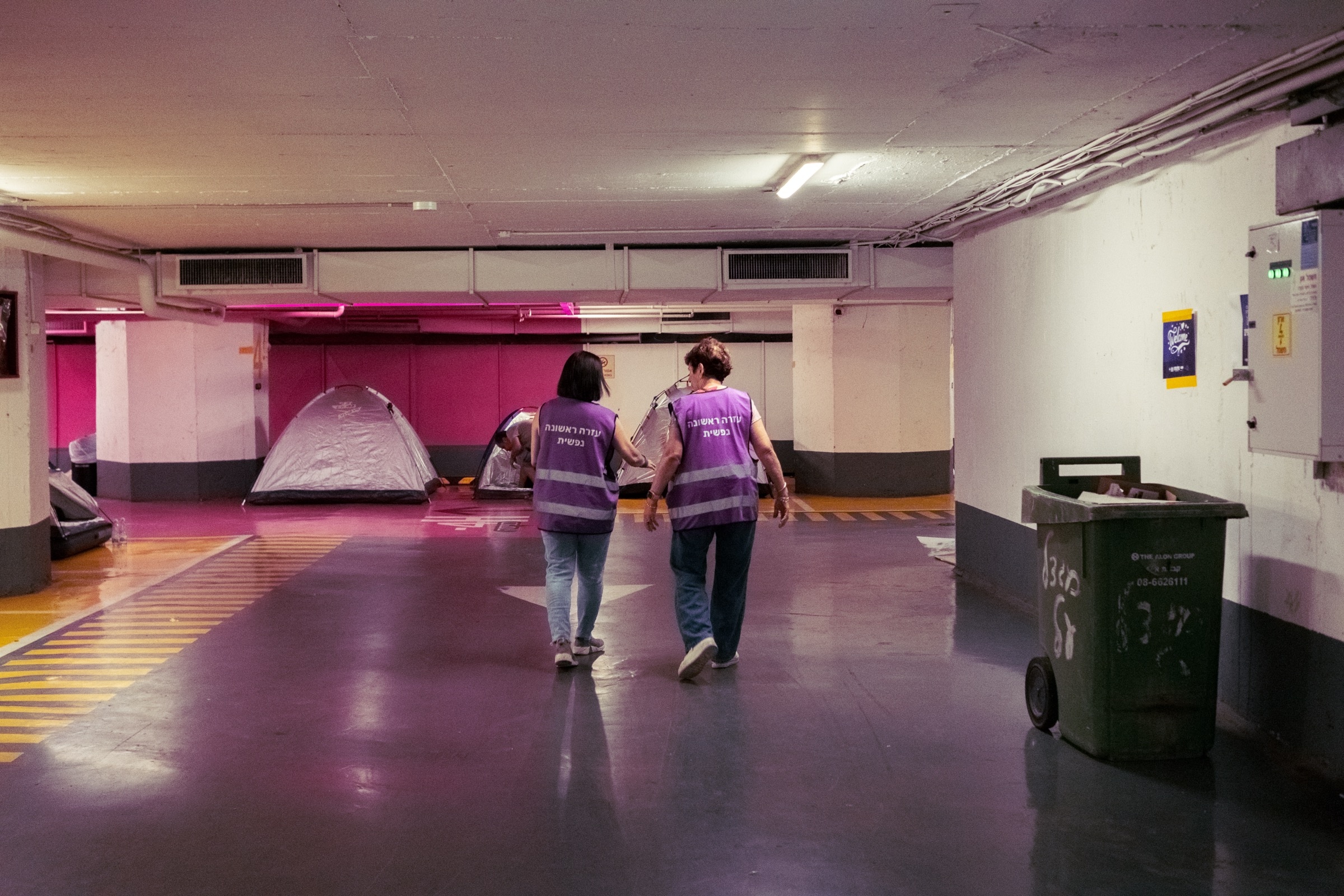 Two volunteers in purple vests walk through a parking garage past tents and a dumpster