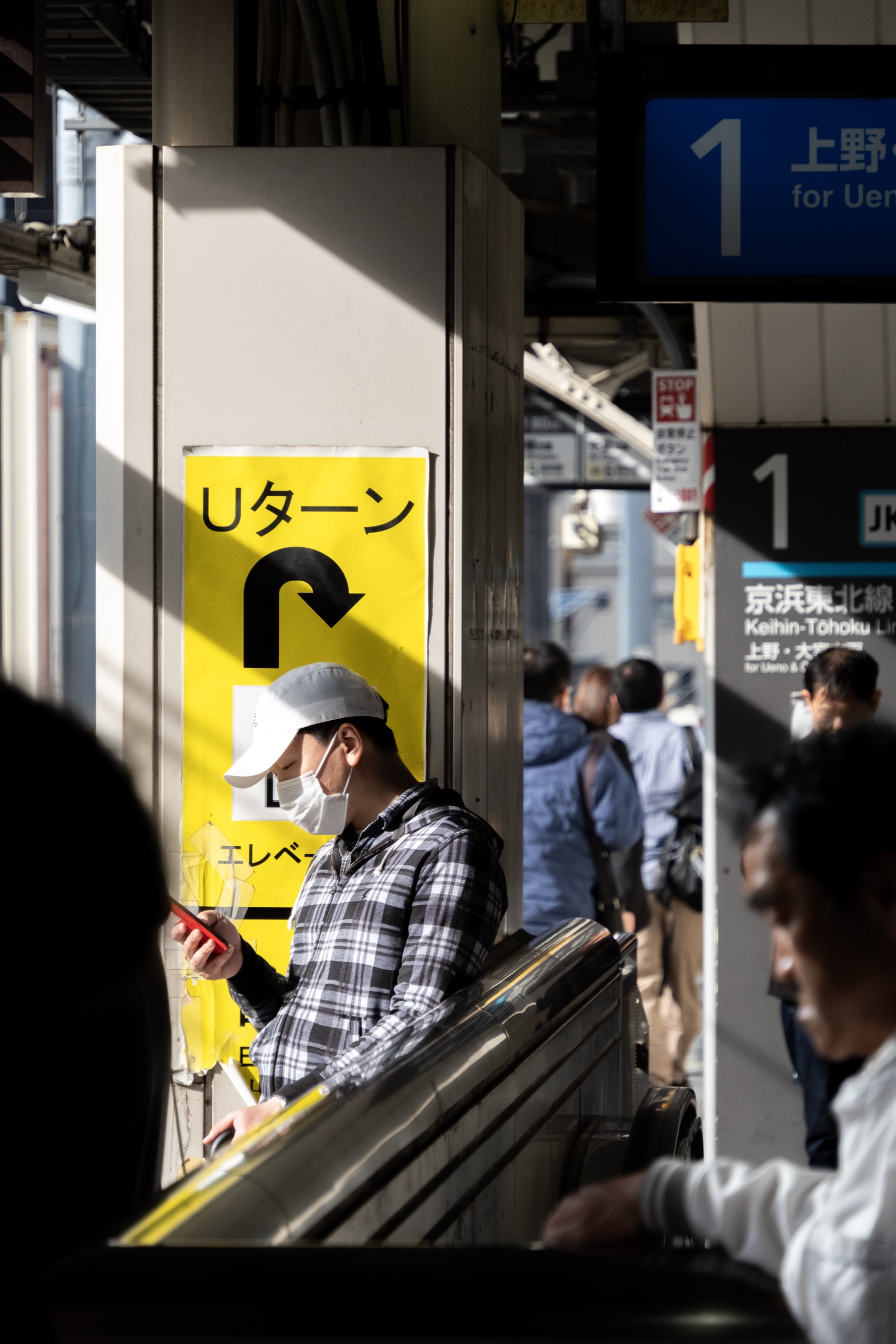 A man in a plaid shirt and mask checks his phone on a train platform beside a yellow U-turn sign, bound for Ueno