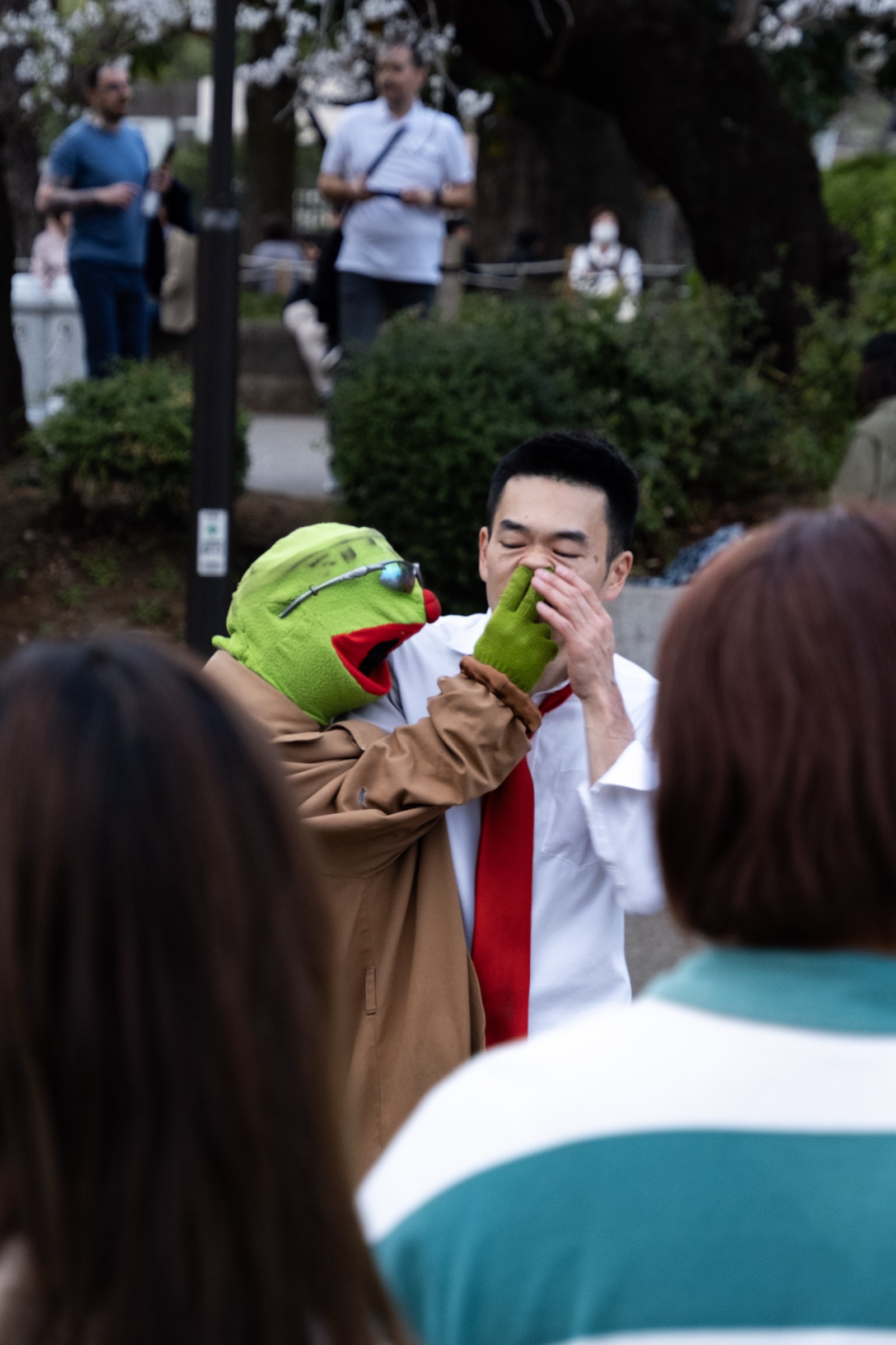 A street performer with a Kermit-the-Frog puppet pinches its green hand against his nose as a crowd watches in a park