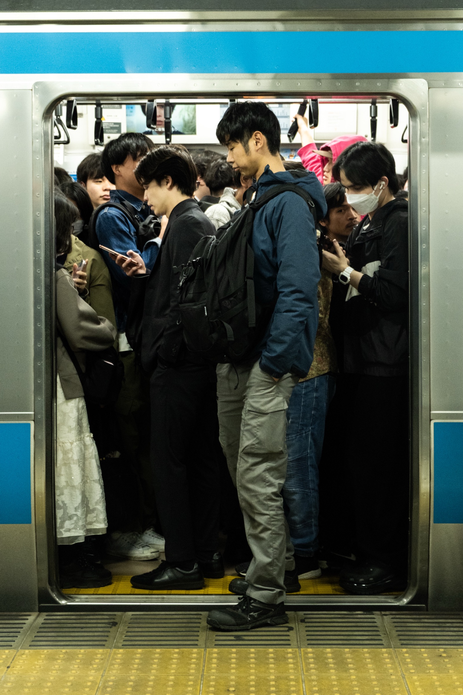 Commuters stand packed shoulder to shoulder in a train car with blue-striped doors, one man's foot on the yellow platform edge