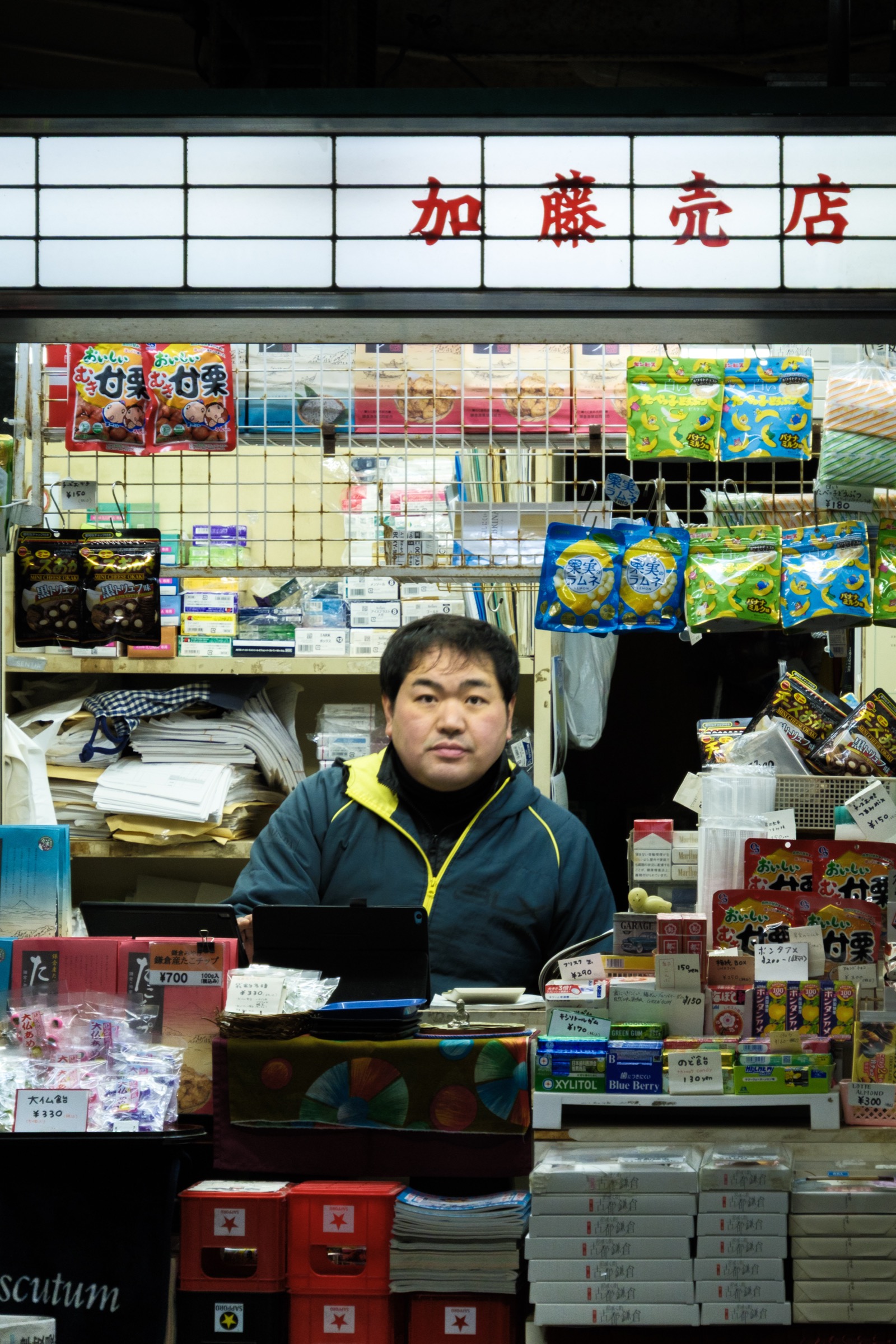 A vendor in a yellow-trimmed jacket stands behind the counter of a small station kiosk stocked with snacks and souvenirs