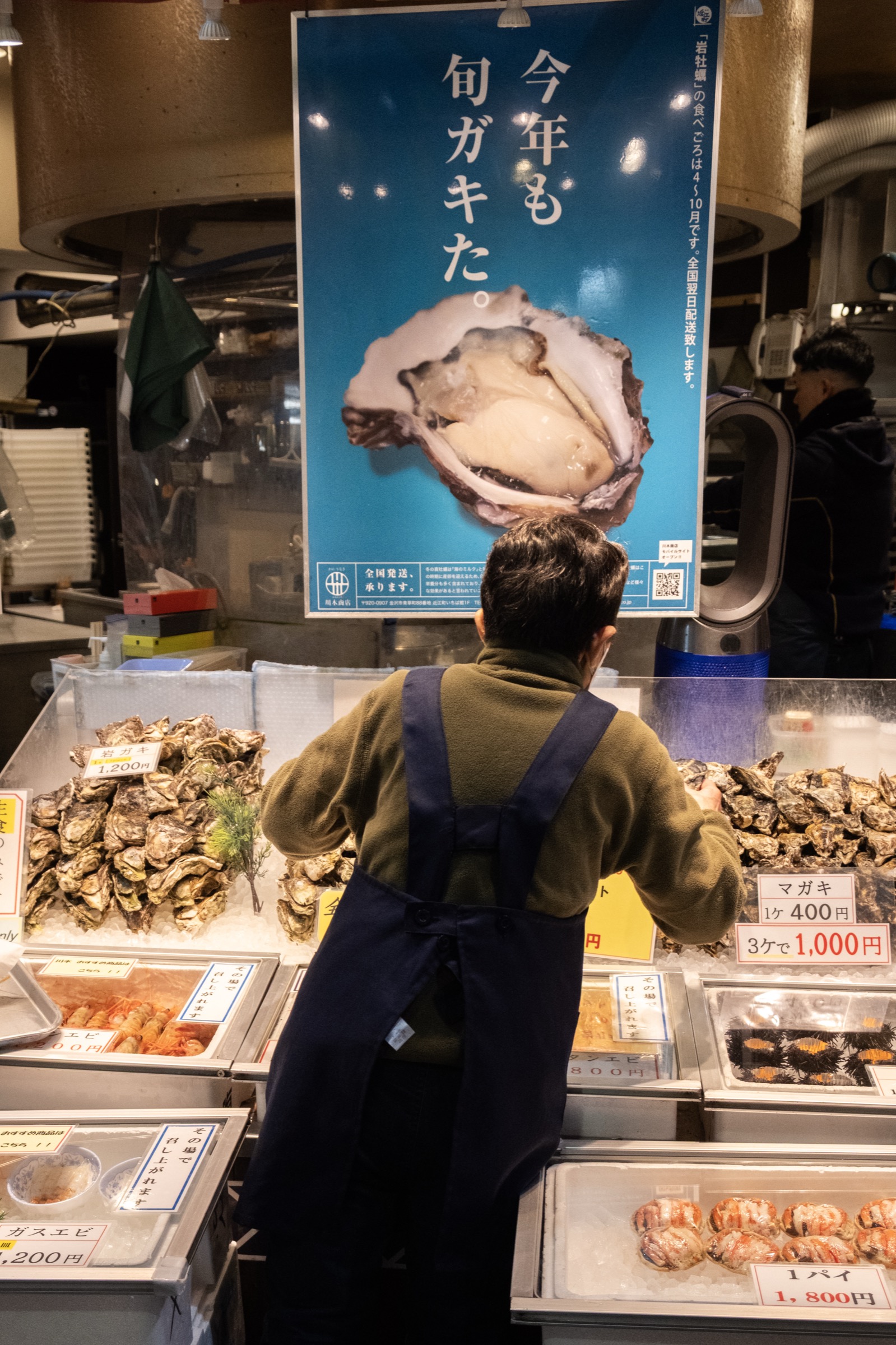 A vendor in an apron stands behind trays of oysters and seafood beneath a large blue poster advertising seasonal oysters