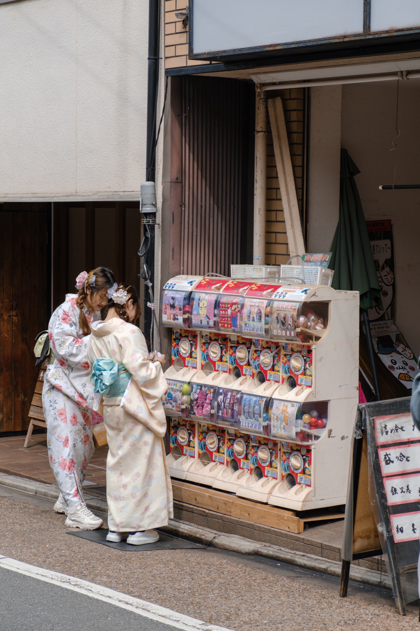 Two young women in floral kimonos browse a row of gachapon capsule-toy machines on a street