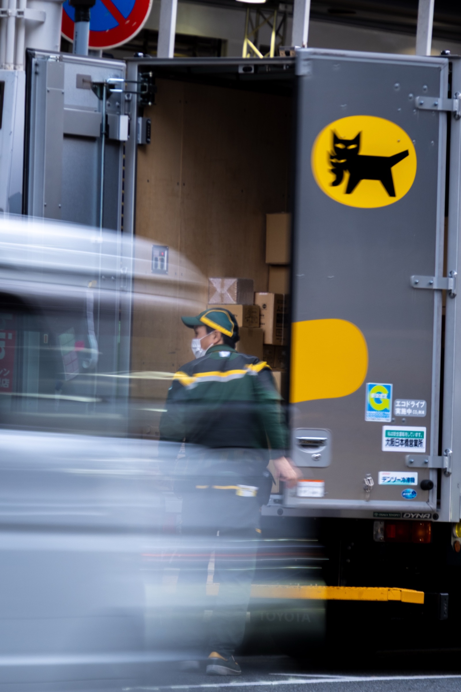 A Yamato Transport driver loads packages from an open delivery truck with the black-cat logo, traffic blurred in the foreground