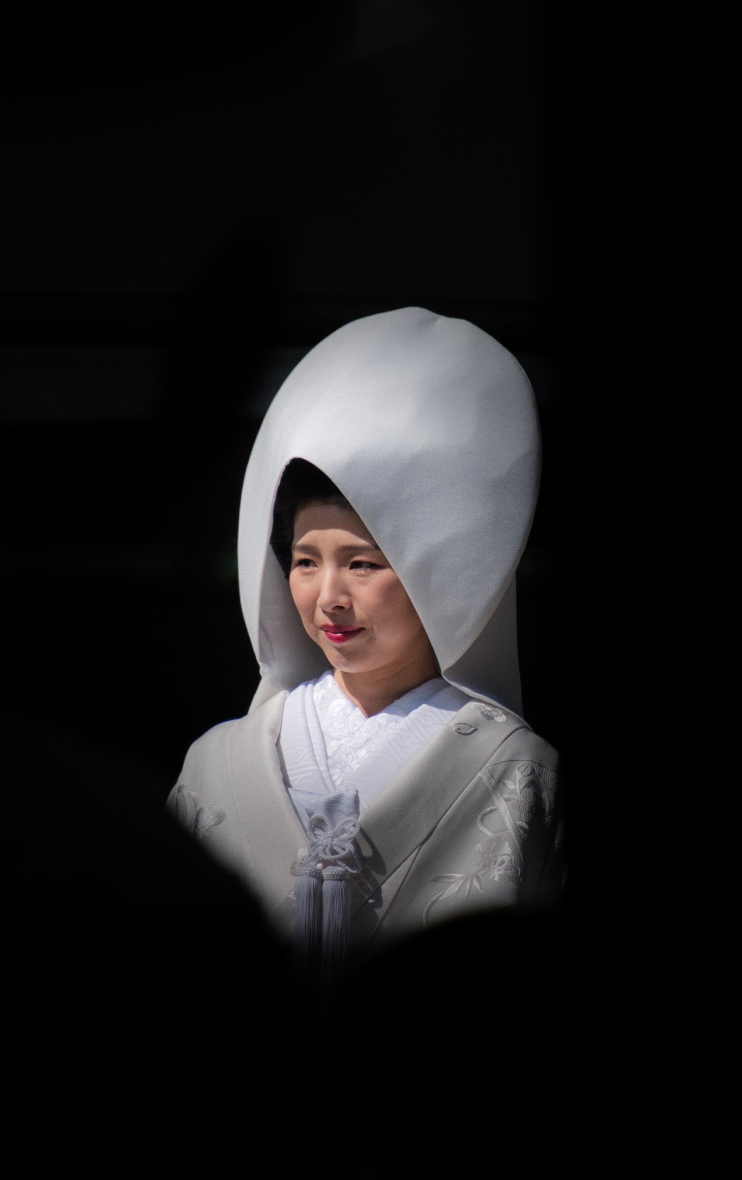 A bride in a white watabōshi hood and shiromuku kimono smiles softly, framed by darkness
