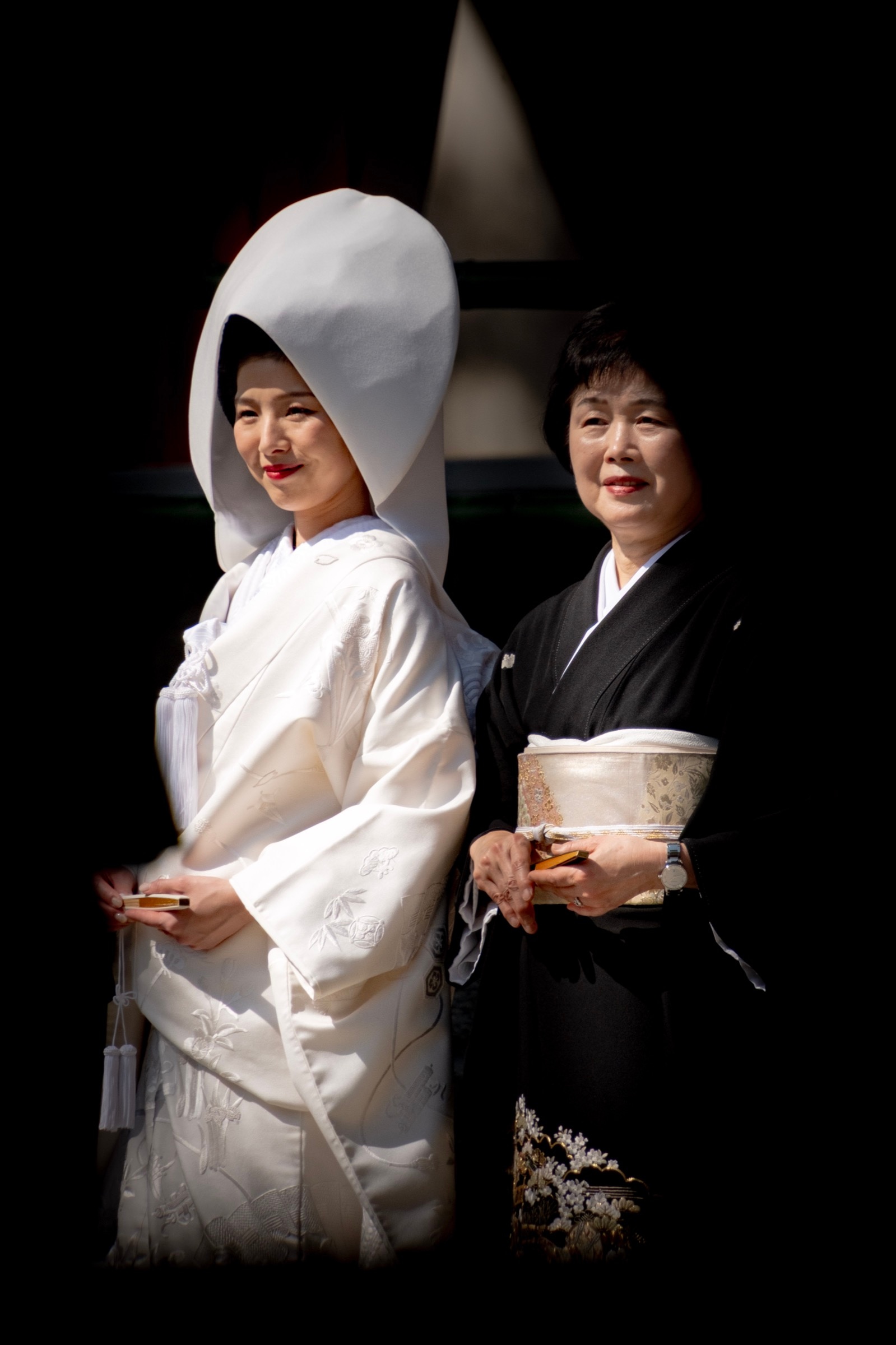 A bride in a white shiromuku stands beside an older woman in a black tomesode kimono with a gold obi, both holding fans