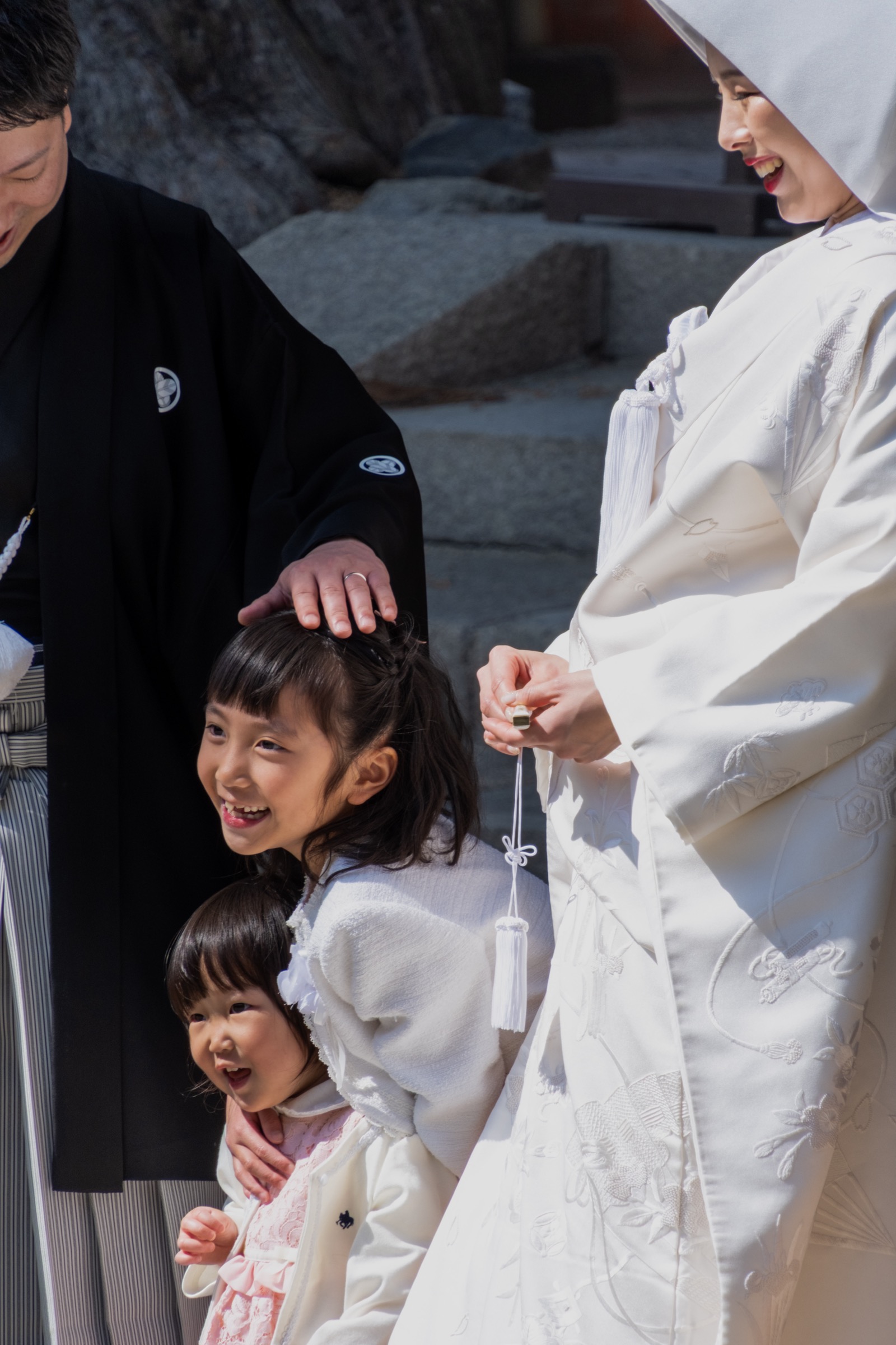 Two grinning young girls peek out from between a groom in a black montsuki and a bride in white shiromuku