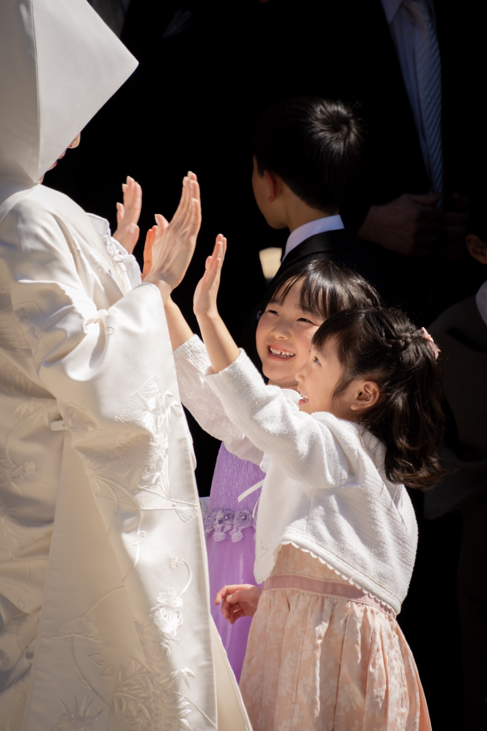 A smiling girl in a pink dress reaches up to high-five a bride in white shiromuku