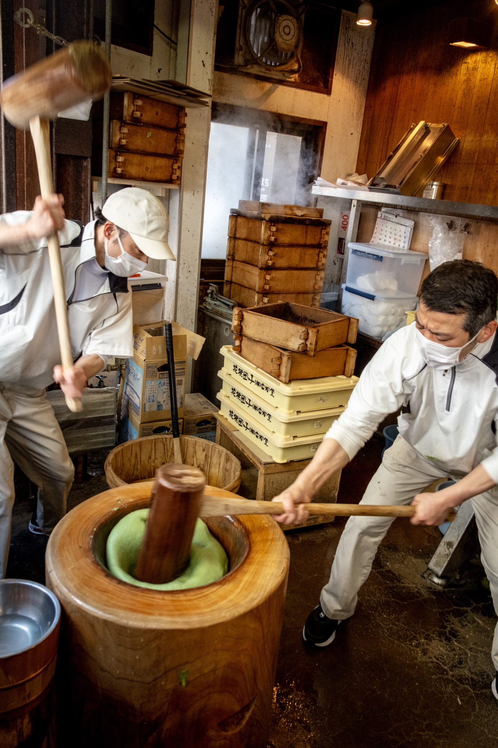 Two men in white uniforms pound green mochi with wooden mallets in a large wooden mortar, steam rising from stacked boxes behind
