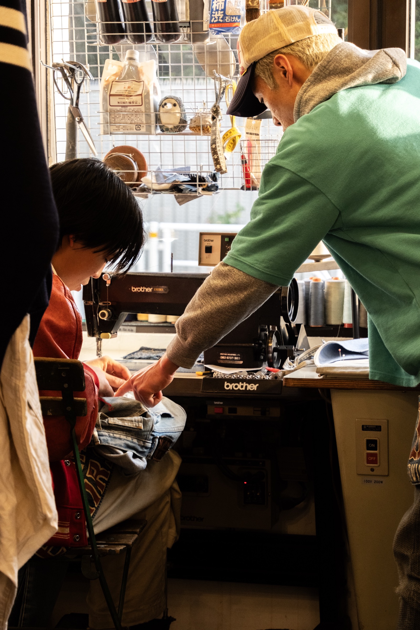 A man in a green sweatshirt and cap hands fabric to a woman seated at a sewing machine in a small workshop